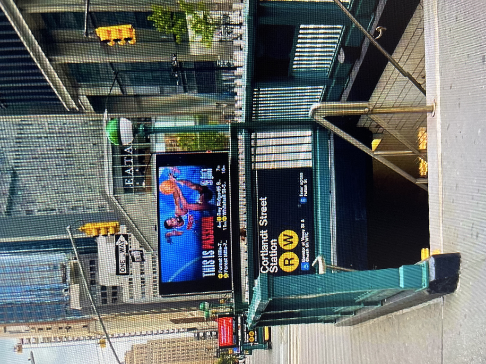 An outdoor daytime shot captures a New York City subway entrance with the sign "Cortlandt Street Station R W". The entrance is surrounded by green railings and a metal fence. Above the entrance, a digital billboard displays an advertisement featuring a basketball player. In the background, tall buildings with glass facades dominate the cityscape. Traffic lights are visible, one yellow and one red, indicating traffic signals. The overall scene depicts a bustling urban environment with elements of transportation and commerce.
