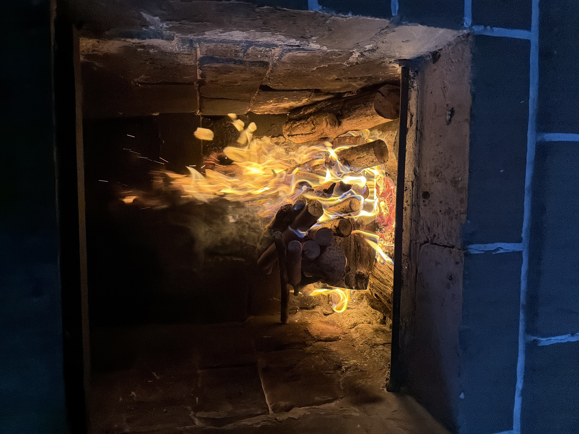 The image shows a close-up view of a fireplace with a fire burning. The fireplace is constructed of dark bricks and stone. The fire is bright and active, with flames reaching upwards and sparks flying. Several logs are visible in the fire, some burning intensely and others still intact. Smoke is rising from the fire, creating a hazy effect. The overall impression is one of warmth and coziness. The image does not contain any information about location or people.