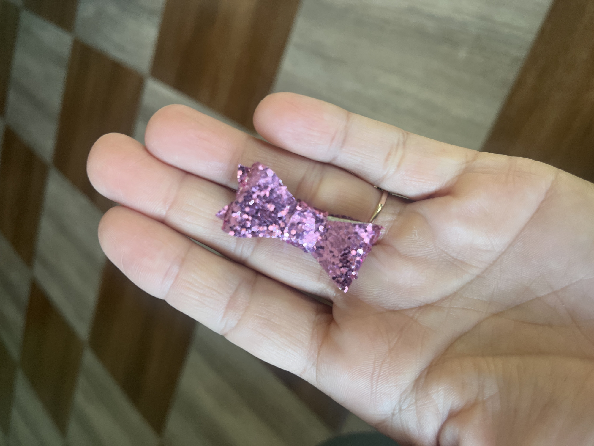A close-up shot focuses on a person's hand holding a small, pink, glittery bow. The hand is lightly cupped, with the bow positioned in the palm area. The bow appears to be made of felt or a similar material, covered in fine pink glitter. A thin gold ring is visible on the ring finger of the hand. The background consists of a tiled floor with alternating brown and beige squares, suggesting an indoor setting. No specific location in Tarnivtsi, Ukraine, can be identified from the visual information provided.