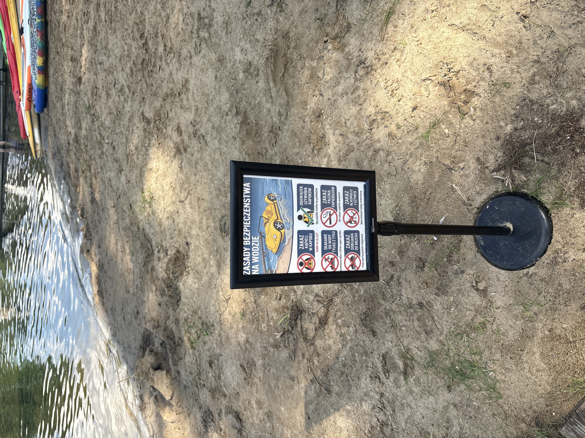 The image shows a sandy beach area next to a body of water. A sign is prominently displayed in the center of the frame, mounted on a black pole. The sign has a black frame and features a graphic of a yellow car driving on water, alongside text written in Polish. The text appears to be safety regulations related to water activities. The sign is positioned on the sand, with a few sparse green blades of grass growing in the foreground. In the background, the water's surface is visible, reflecting the sky. Further in the background, on the water's edge, several colorful paddleboards or kayaks are stacked. The overall environment suggests an outdoor recreational area, possibly a lake or riverfront.