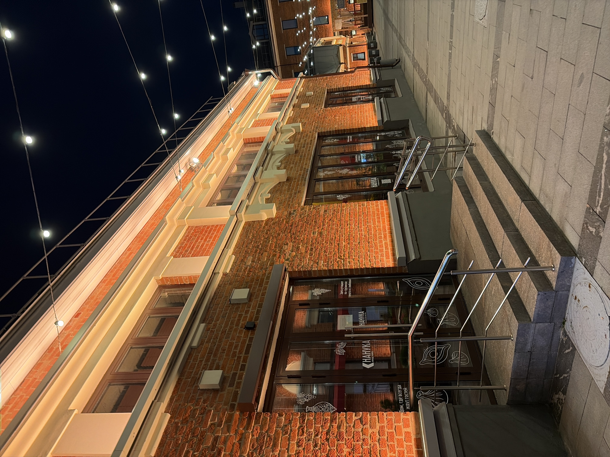 The image depicts an exterior view of a brick building at night, likely a commercial establishment, in Moscow, Russia. The building is adorned with warm, indirect lighting along its upper facade and around the windows. A string of decorative lights stretches across the dark sky above. In the foreground, there are steps leading up to glass double doors, which have signs with Cyrillic text and graphical elements of grapes and other produce. The sidewalk is paved with stone slabs. In the background, another building with similar architectural features and string lights is visible, suggesting a street or courtyard setting. The scene is tranquil and illuminated, characteristic of a European city at night.