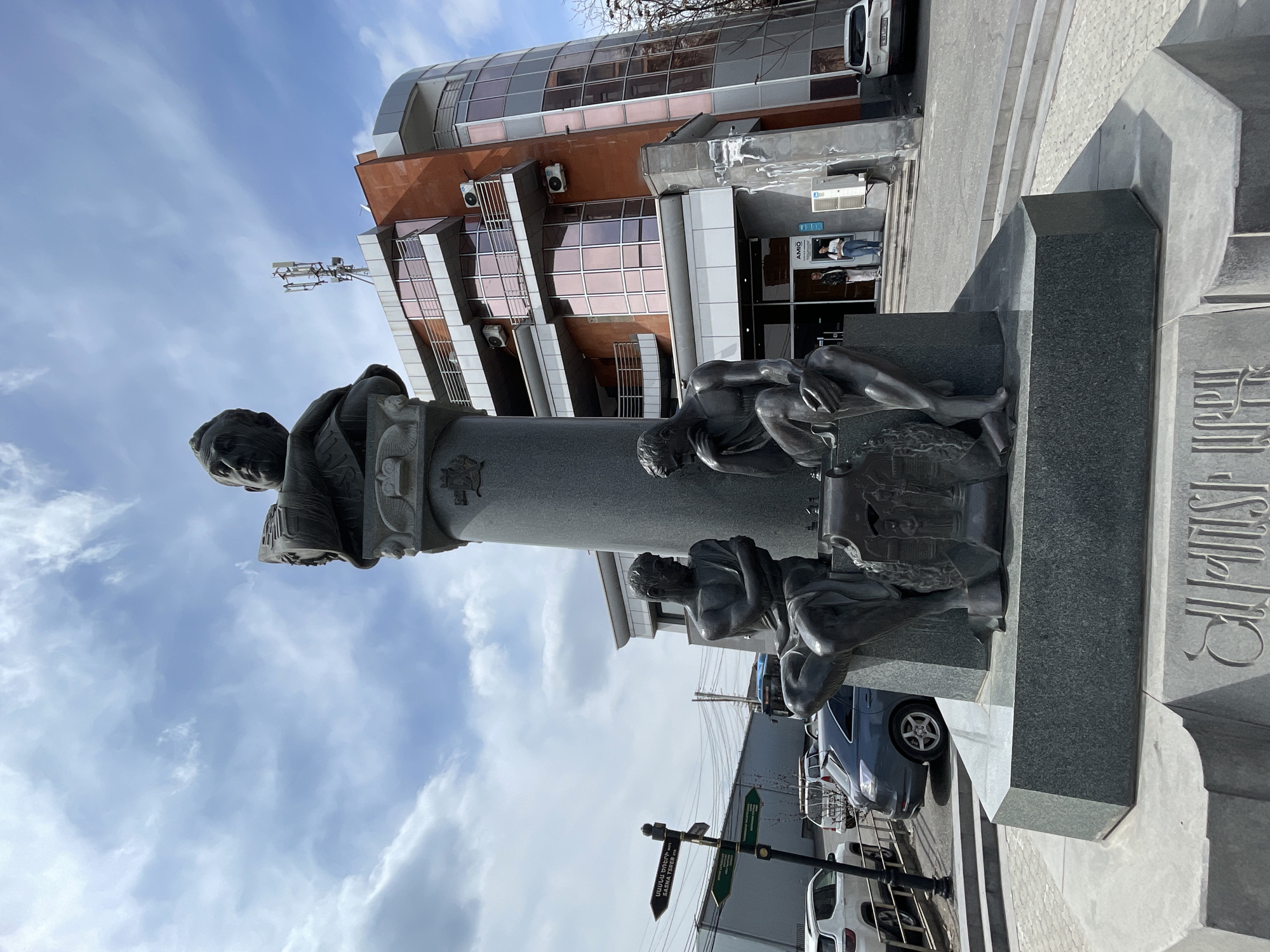 The image depicts the monument to Hovhannes Tumanyan in Yerevan, Armenia. The monument comprises a bust of Tumanyan atop a column, with sculpted figures at the base. The figures appear to be in various poses, some kneeling or leaning. The background shows a modern building with mirrored windows and what appear to be office spaces. Street signs are visible near the base of the monument, along with vehicles, indicating that the monument is situated in a public space near a road. The sky is partly cloudy. The monument appears to be made of stone or concrete.