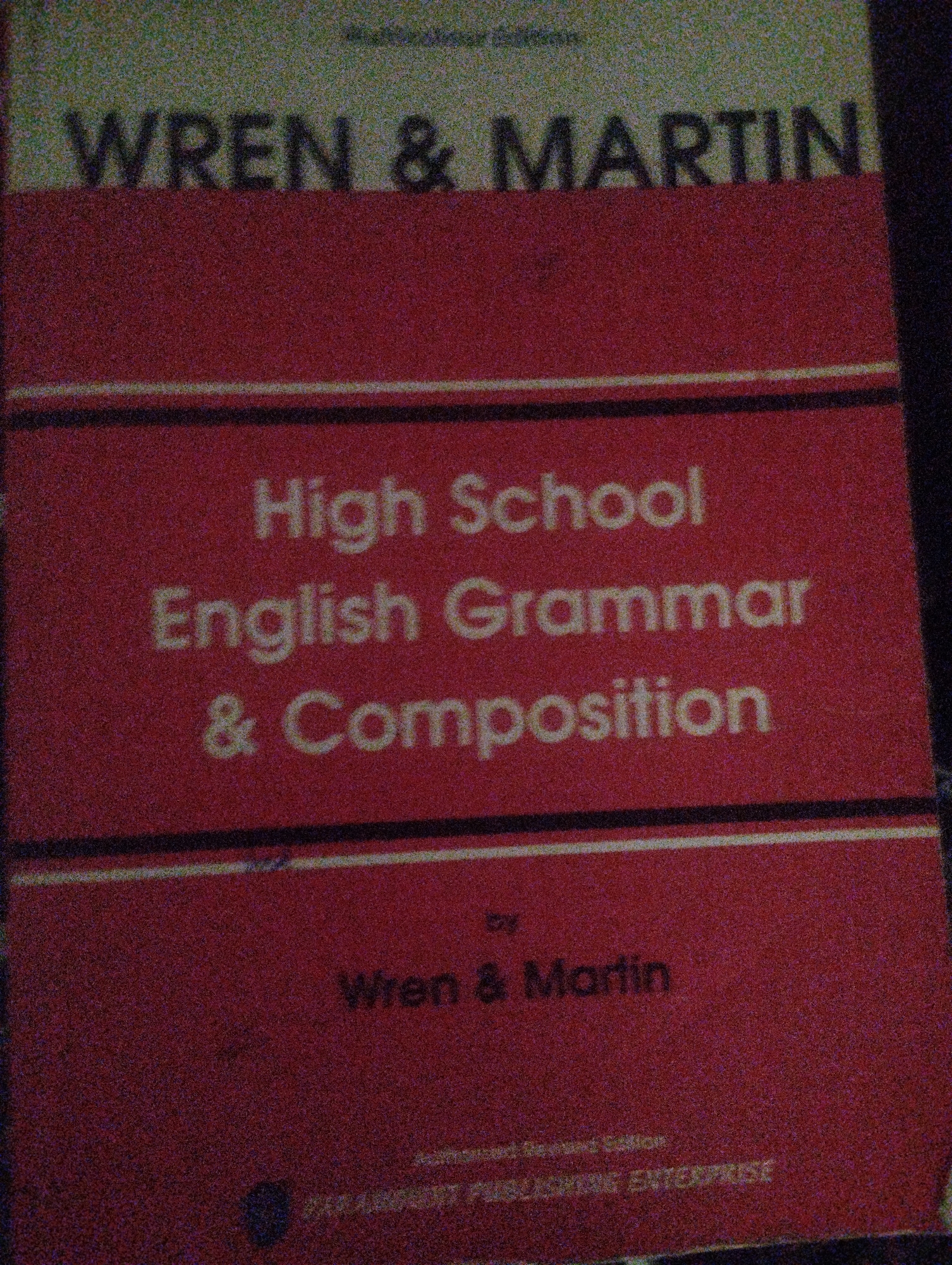 This is a close-up, slightly grainy photograph of the cover of a book titled "WREN & MARTIN High School English Grammar & Composition". The cover features a red background with white text. The title is prominently displayed in large, bold white letters. Below the main title, the words "by Wren & Martin" appear in smaller white text. At the very bottom of the cover, a publisher's name is visible, though slightly obscured by the grain and angle of the photo. The overall impression is of an educational textbook. There are no people, animals, or specific settings visible in the image, only the book cover itself. The lighting suggests it was taken indoors, possibly at night, given the quality of the image.