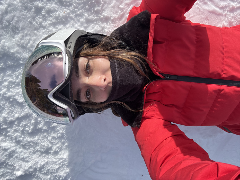 A person wearing a red snowsuit, ski goggles, and a black neck gaiter lies in the snow. The goggles reflect trees and sky. The person looks directly at the camera. The zipper of the snowsuit is partially open. The snow is bright and white.