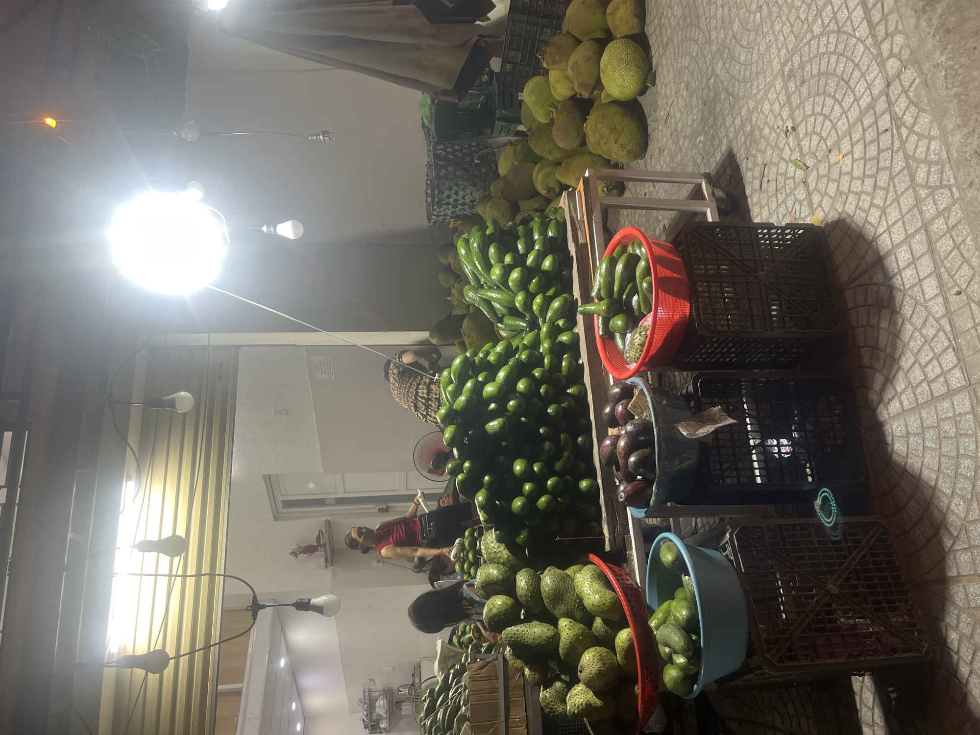 This is an exterior night shot of a fruit stand located on a tiled sidewalk, possibly in the city of Đà Nẵng, Vietnam. A woman in a red shirt and dark pants is standing behind a table laden with fruits, interacting with a person whose back is to the camera. Several large piles of avocados are prominent. To the left, there are also piles of soursop and other green fruits. In the background, more fruits, possibly jackfruit, are visible, along with two black plastic crates and an umbrella. Several light bulbs are suspended overhead, illuminating the scene. The ground is paved with circular patterned tiles.