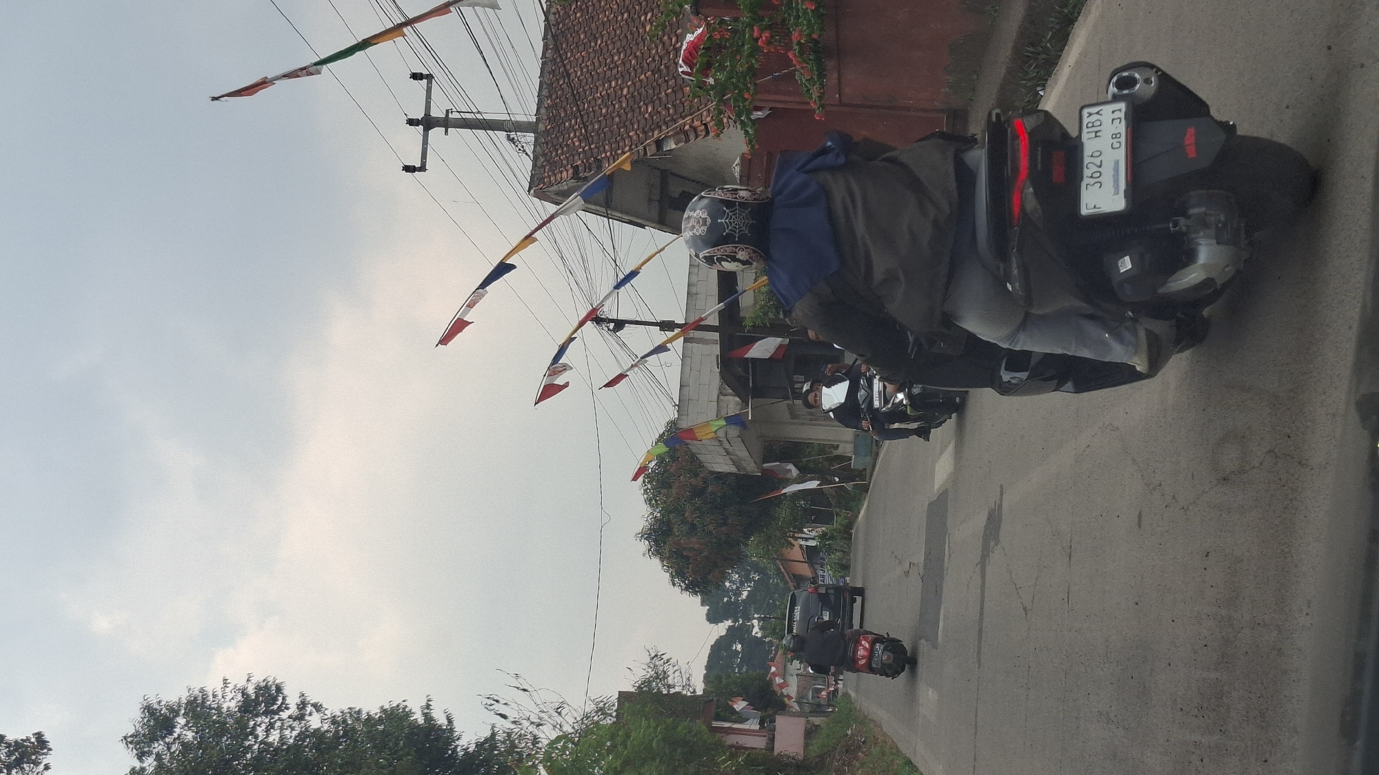 The image depicts a street scene in Sirnagalih, Indonesia, on an overcast day. The perspective is from a vehicle, looking forward. In the foreground, a person is riding a black scooter. This rider is wearing a helmet with a spiderweb design and a dark jacket over light-colored pants. The scooter has a license plate that reads "F 3626 HBX GB 31".
Further down the road, two other motorcycles are visible. One is a red scooter with a rider in black, and another is a dark motorcycle with a rider wearing a white mask. A dark SUV is also present in the traffic.
The street is paved with asphalt and shows signs of wear and tear, including cracks. On the sides of the road, there are buildings with red roofs and a power pole with multiple utility wires. Colorful festive flags, consisting of red, white, blue, and yellow stripes, are strung along poles on both sides of the street, suggesting a local celebration or event. Lush green trees and foliage are also visible along the roadside. The sky is cloudy and pale.