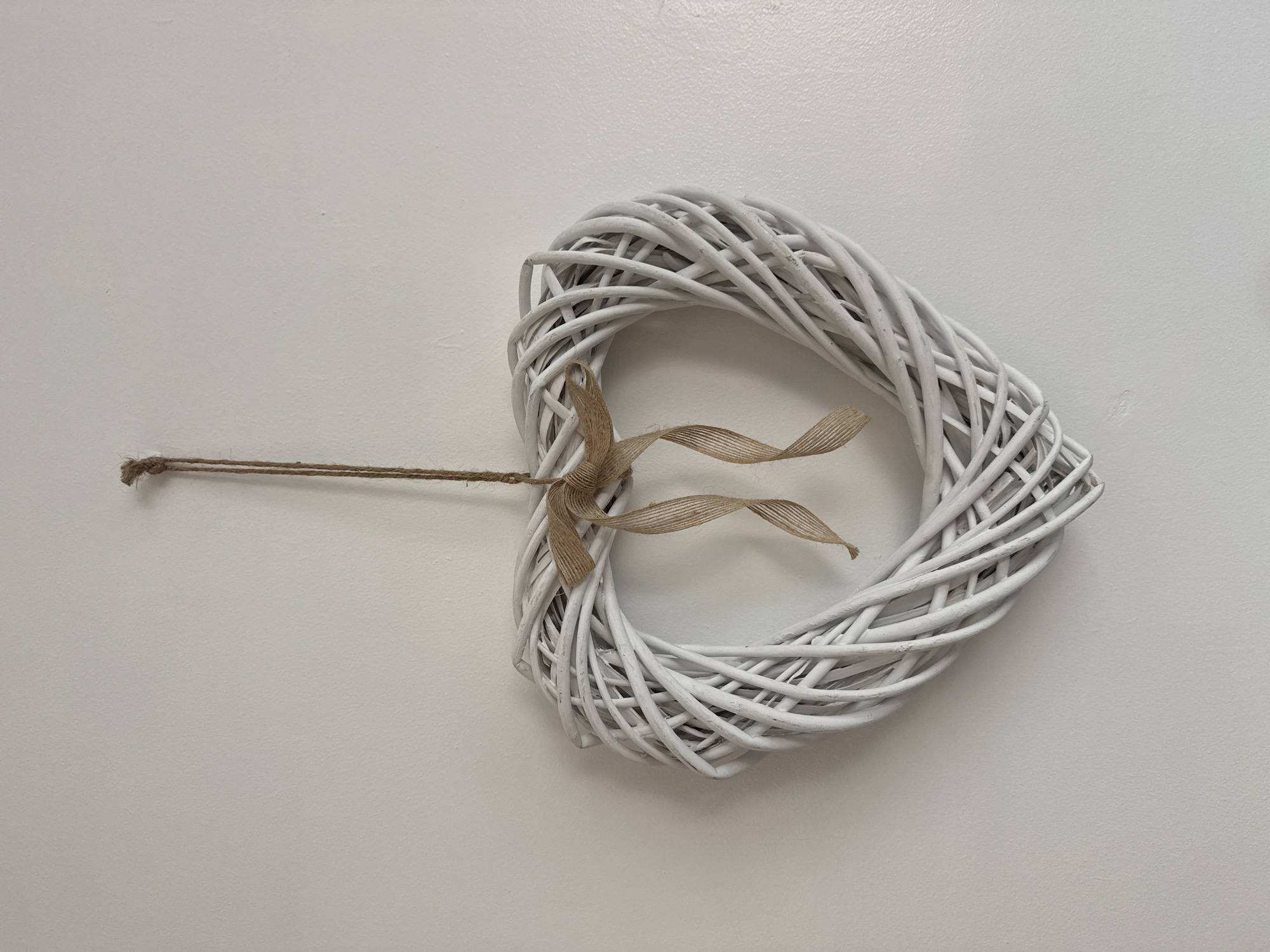 A white wicker heart-shaped wreath hangs from a single piece of twine against a plain white wall. The twine is knotted at the top and secured to the wall. A small, tan burlap bow is tied around the top curve of the heart, with two ribbon-like strands hanging down from it. The wreath is made of interwoven branches, painted white, with some visible texture from the material. The wall appears to be painted plaster. There are no people or other objects visible in the image. The scene is likely indoors, possibly in a home or a shop in Darlington, United Kingdom.