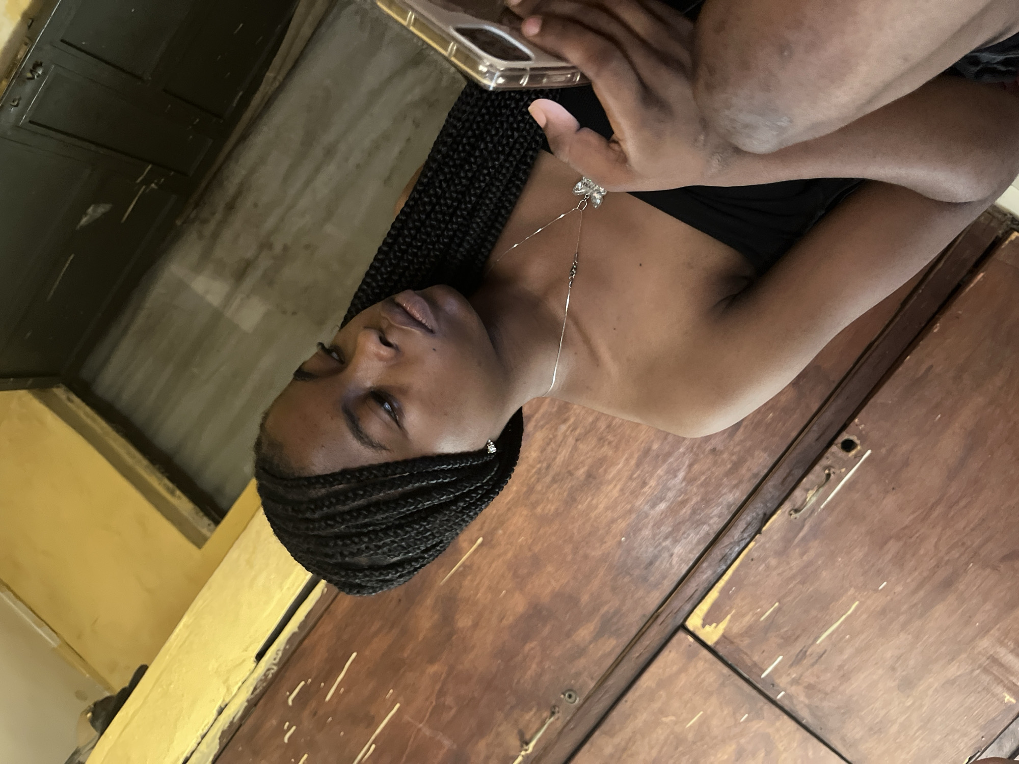 A young woman with braided hair is sitting and looking at her phone. She is wearing a black strapless top and a necklace with a butterfly pendant. She is sitting in front of a wooden door and a yellow wall. The environment appears to be indoors.