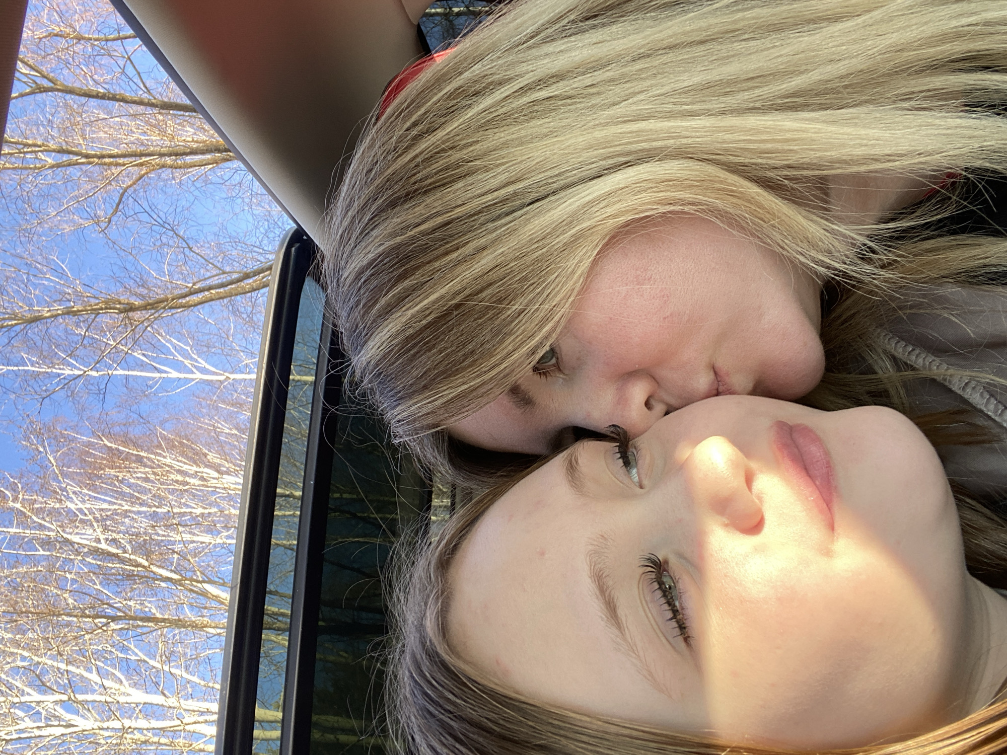 An intimate moment is captured inside a vehicle, featuring two individuals, likely two females, in close embrace. The image is oriented horizontally, with the subjects' faces close together. An individual with long, bright blonde hair is seen leaning down to gently kiss the forehead or temple of another, younger individual who has lighter brown or blonde hair and is looking upwards with open eyes.
The scene appears to be set inside a car with an open window, as indicated by the visible dark frame of the car window and a portion of the car's interior lining on the right and top. Beyond the vehicle, a clear blue sky serves as the backdrop for numerous bare, light-colored tree branches, characteristic of deciduous trees in late winter or early spring. These trees strongly resemble birch trees, which are common in the implied location of Chaikovsky, Russia.
The bright, natural sunlight illuminates the subjects, casting a prominent highlight across the lower person's face, emphasizing their features and eyelashes. The sunlight also brightens the blonde hair of the upper individual. The image conveys a sense of warmth, affection, and tenderness, set against a crisp, sunny outdoor environment. No visible text is present.
