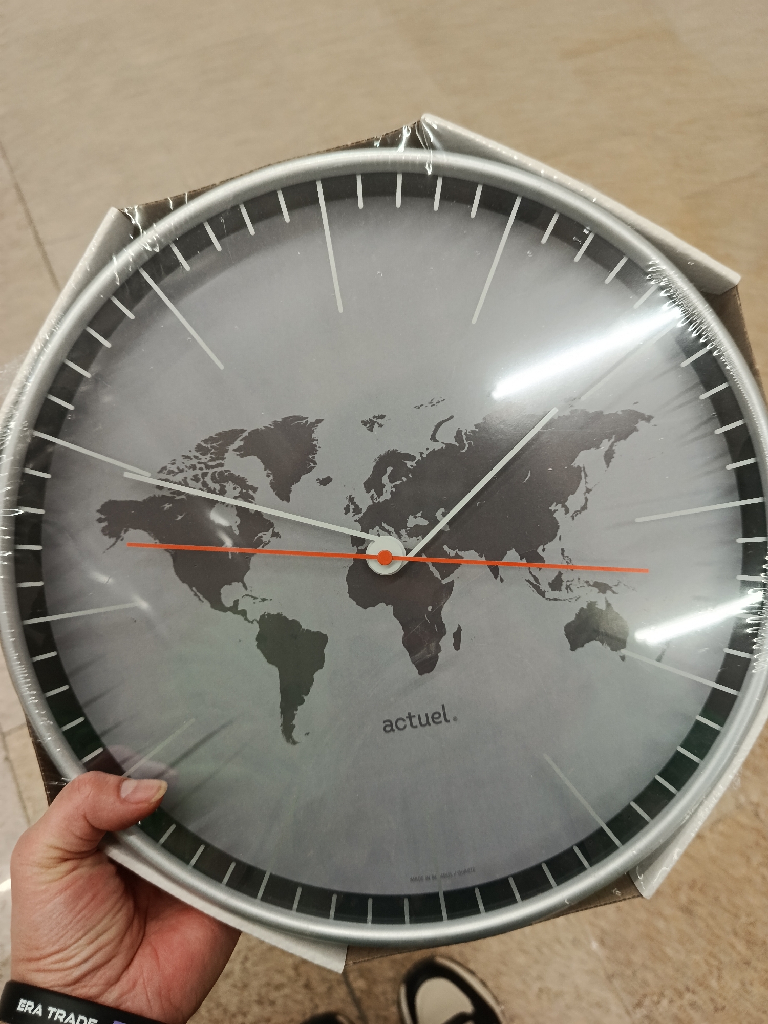 The image shows a clock with a world map design on its face. The clock has a silver frame and a grey background with a dark grey world map. The hour and minute hands are orange. The second hand is white. The clock is still wrapped in plastic. The brand name "actuel." is printed on the clock face near the bottom. The clock is being held by a hand with a black wristband that says "ERA TRADE". A shoe can also be partially seen in the image.
I cannot determine if the location is in Moscow, Russia, as there are no indicators of the specific city or country.