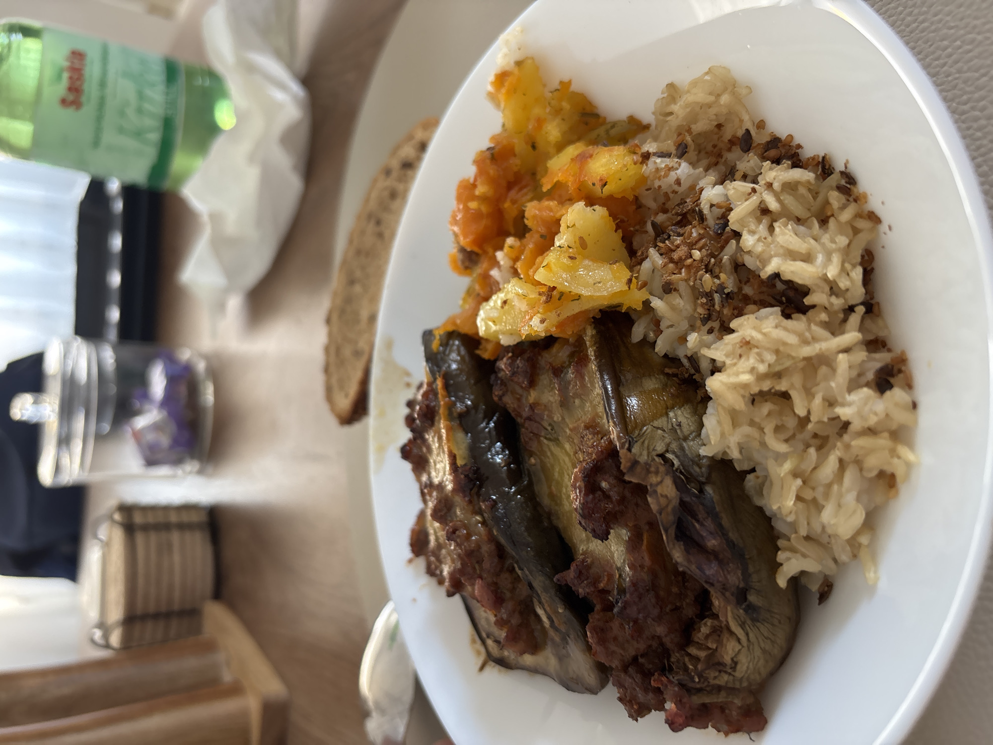 The image shows a close-up of a white plate filled with food, positioned on a wooden table. The meal consists of stuffed eggplant, rice, and what appears to be a mixture of diced vegetables, possibly pumpkin or sweet potato. The stuffed eggplant is dark purple and appears to be filled with a ground meat mixture, topped with melted cheese and baked until slightly browned. The rice is fluffy and light brown, sprinkled with some darker seeds. The diced vegetables are vibrant orange and yellow. In the background, out of focus, are a clear glass jar with a metallic lid, a white paper napkin, a slice of bread, and a green bottle of "Saskia" sparkling water. The setting appears to be a dining area, possibly a restaurant or a home. Based on the "Saskia" brand of water, which is commonly sold in Germany, and the style of the food, the location is likely Viernheim, Germany. No people are visible in the frame.