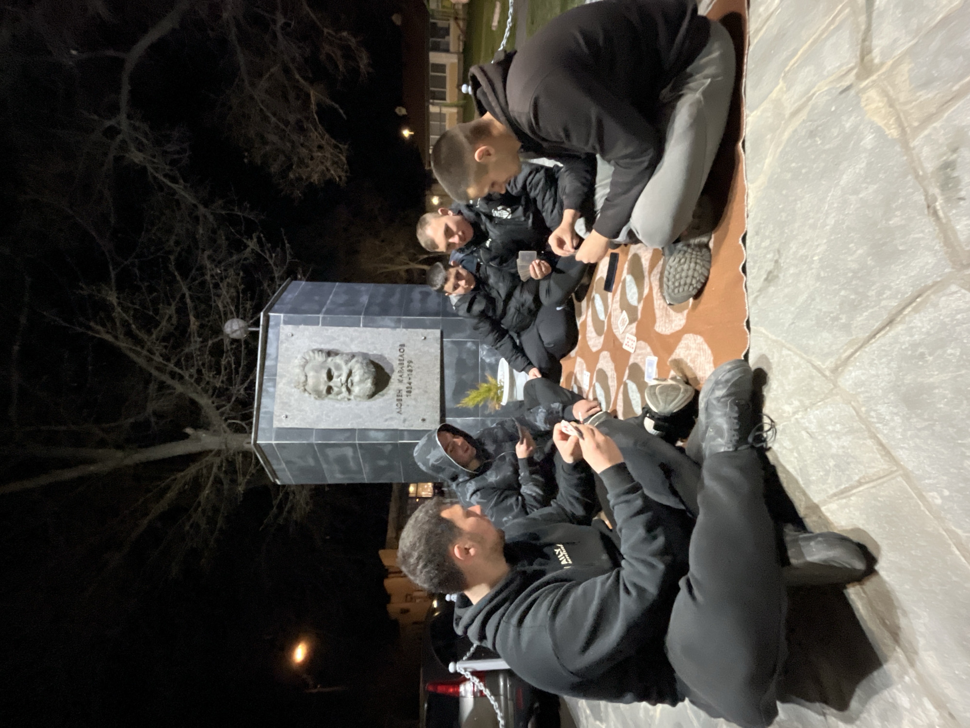 A group of six individuals are gathered at night in the city of Karavelovo, Bulgaria, sitting on a blanket on the ground in front of the monument of Lyuben Karavelov. The individuals are playing cards. The monument itself is made of grey stone and features a bust of Karavelov. There is a tree to the left of the monument. It is dark. A black car is parked to the right.