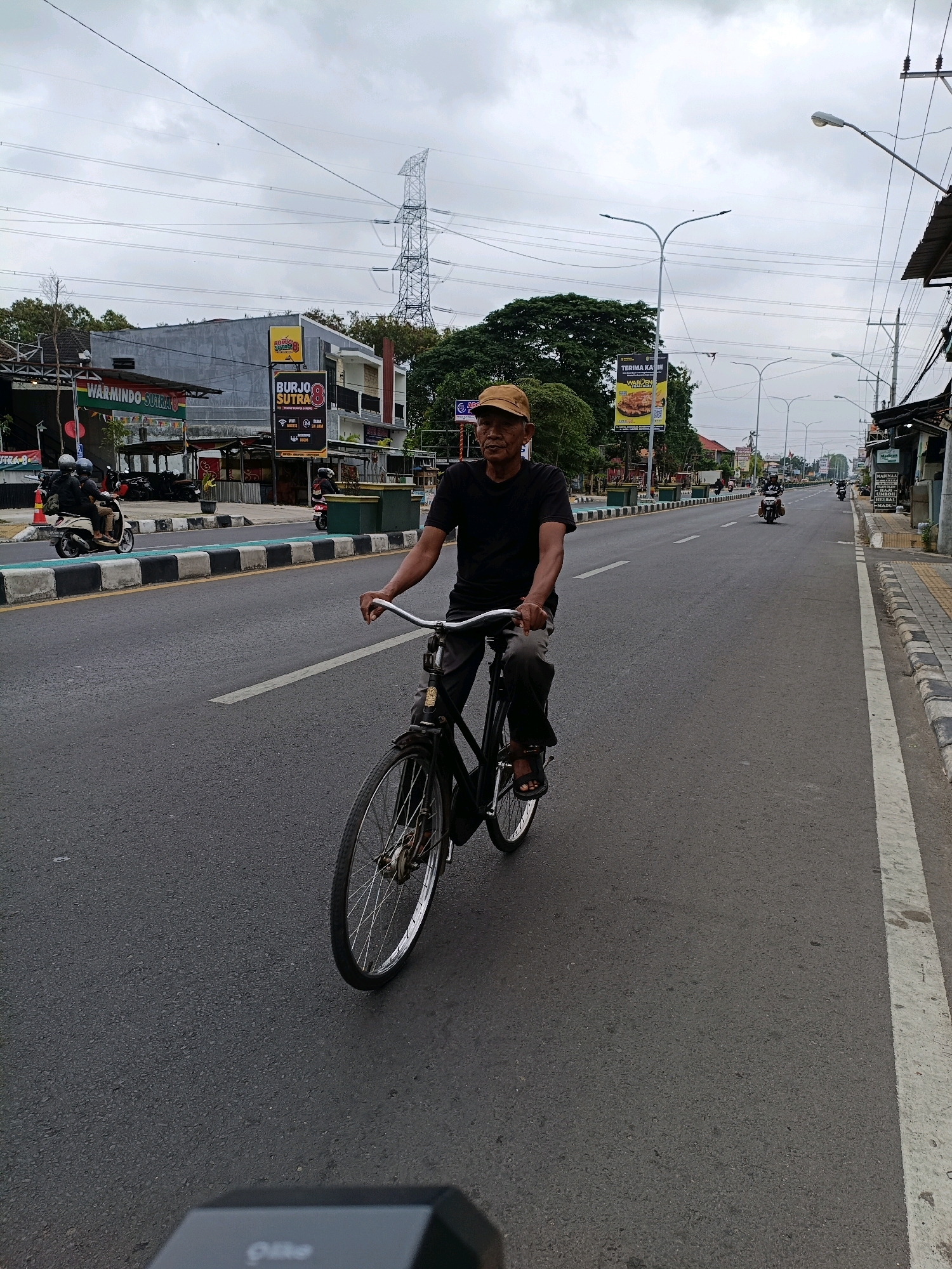 An elderly man with a brown cap and dark t-shirt rides a black bicycle down a paved road in Sewon, Indonesia. The man is positioned in the center of the frame, facing left, with his left hand on the handlebars and his right arm bent. His legs are extended, with his left foot on the pedal. The bicycle has a silver front wheel with spokes and a dark-colored frame. In the background, there are various buildings and businesses lining the road, including one with a large yellow sign that reads "BURJO SUTRA 8". There are also several motorcycles and scooters on the road, some parked and some in motion. Overhead, power lines crisscross the sky, and a tall electrical tower is visible in the distance. The sky is overcast with clouds, suggesting a slightly gloomy day. The road is marked with white lines and has a green bicycle lane along the left side. The overall scene depicts a typical streetscape in a populated urban or semi-urban area of Indonesia.
