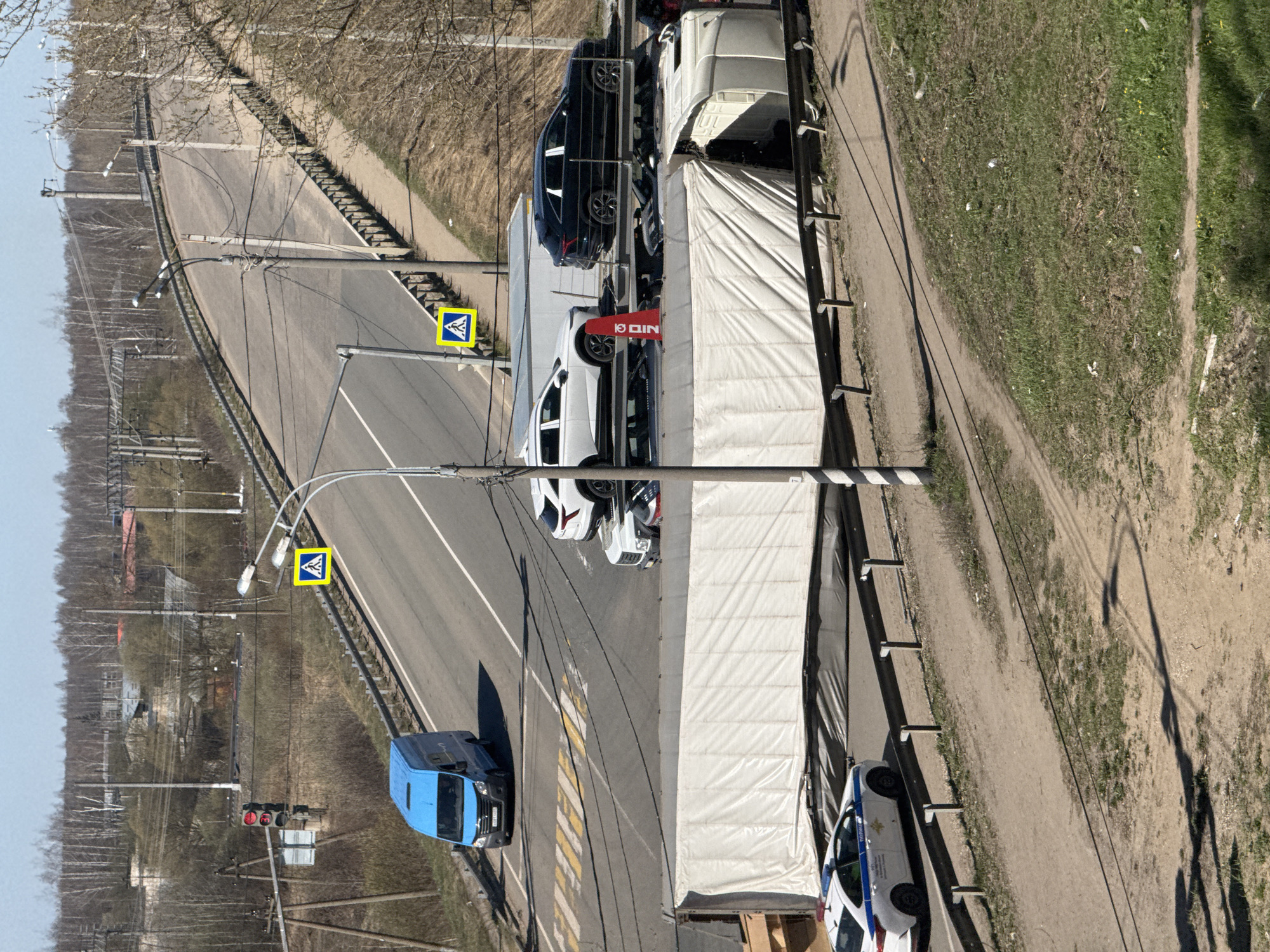 An elevated view shows a street scene in Ново-Никольское, Russia. A blue van is on the left side of the road. Vehicles are parked along the right side of the road, with a large truck partially covered by a white tarp. Pedestrian crossing signs are visible on lampposts. A row of bare trees lines the road to the left, and a grassy area is on the right. Traffic signals are visible further down the road to the left.