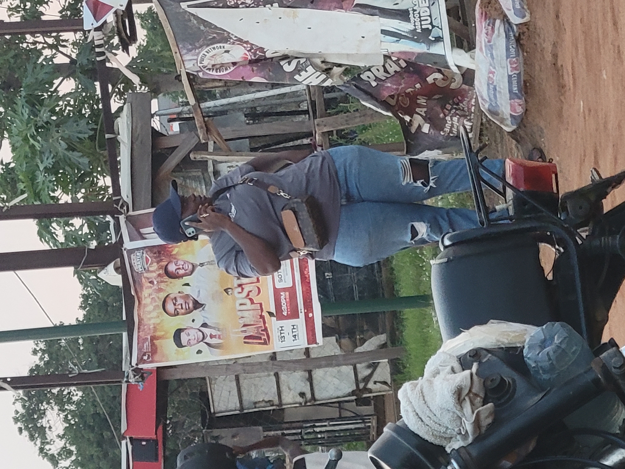 The image captures a scene likely in Suleja, Nigeria, given the "TANGOTE" (Dangote) cement bag. An individual wearing a cap and a sling bag is on a phone call, standing near a poster titled "LAMPST" featuring three men's faces, with event dates 13th and 14th at 4:30 PM and the words "PRAYER," "SOT," and "AM." There's also another sign with "PUTER NETWORK WORD" visible above other banners. A motorcycle is prominently featured in the foreground.