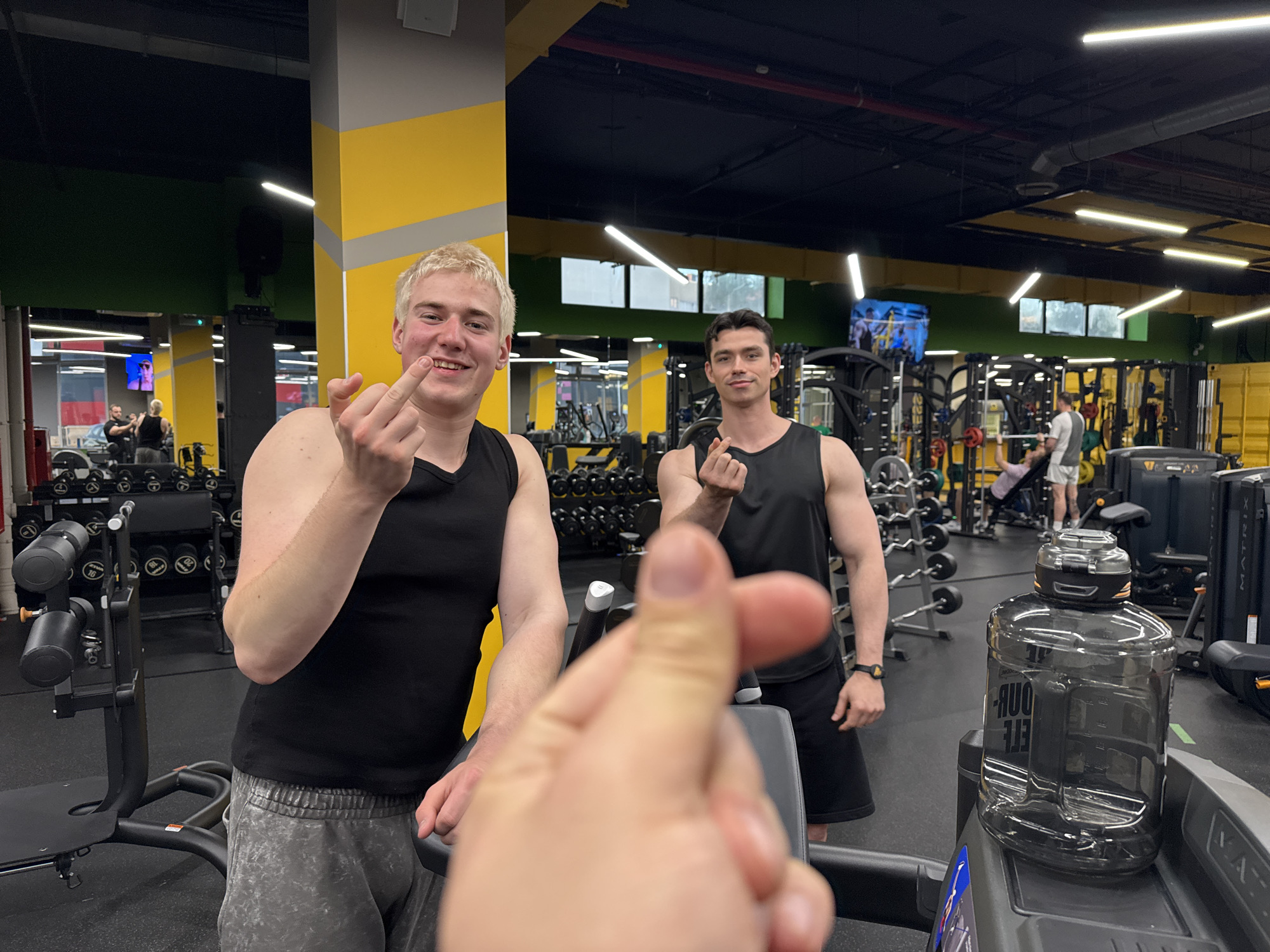 This image captures a vibrant scene inside a modern gym, focusing on three young men posing for a photo. In the foreground, a blurred hand, likely belonging to the photographer, makes a "finger heart" gesture. To the left, a blonde-haired man in a black tank top and grey sweatpants smiles broadly while playfully extending a middle finger towards the camera. Beside him, a dark-haired man, also in a black tank top and black shorts, offers a faint smile and forms a "finger heart" with his left hand.
The gym environment is well-lit with linear fluorescent fixtures on a black ceiling, complemented by yellow and grey structural pillars. Various exercise equipment, including weight racks, dumbbells, and cardio machines, fills the background. Several other individuals are visible throughout the gym, some actively exercising on machines or with free weights, such as a man using a bench press in the mid-ground.
A large, dark-tinted water bottle, appearing to be a gallon-sized container, rests on a treadmill in the right foreground. The text "2.2 LITRE" is faintly visible in reverse on the bottle. The brand "MATRIX" is visible on the console of the treadmill. The overall mood is playful and casual, typical of friends interacting during a gym session.