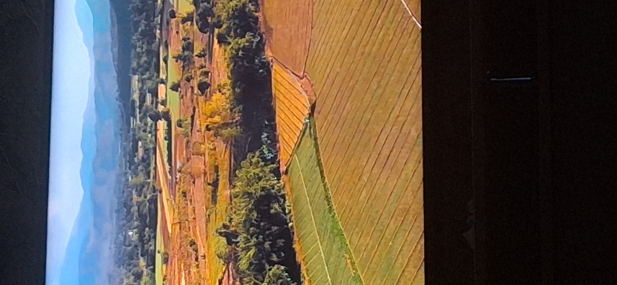 The image displays an aerial view of a rural landscape with agricultural fields. In the foreground, there are several fields, some appearing to be planted with crops, indicated by rows of green and brown vegetation. One field is a distinct bright green, suggesting a recently planted or different type of crop, while others are a muted brown or orange, possibly indicating harvested land or a different stage of growth.
Further into the image, the landscape transitions to areas with trees and scattered buildings, suggesting a village or small town integrated into the agricultural setting. Beyond this, a series of rolling hills or mountains are visible in the distance, layered with soft blue hues, indicating a hazy atmosphere or distance. The sky is a pale, clear blue. The overall impression is one of a vast, cultivated agricultural region. The image does not provide any specific visual cues or landmarks that can be definitively linked to the city of Bila Tserkva, Ukraine.