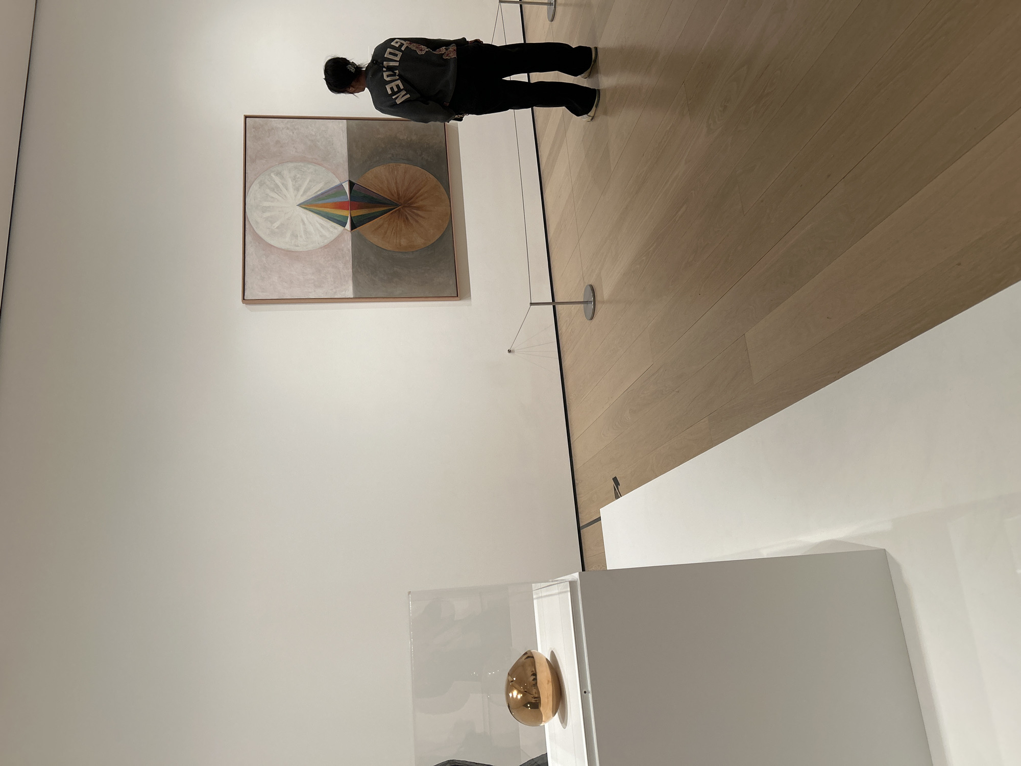 The image shows a museum interior in New York City, United States. A person wearing a dark hoodie with "GOLDEN" printed on the back and dark pants is viewing a large, framed artwork on a white wall. The artwork features geometric shapes, including circles and a segmented design with various colors. A light-colored wood floor extends from the wall with the artwork. A white display case containing a golden, egg-shaped object is visible in the lower portion of the frame. A rope barrier is set up to keep people at a distance from the painting. The lighting is bright and diffused.