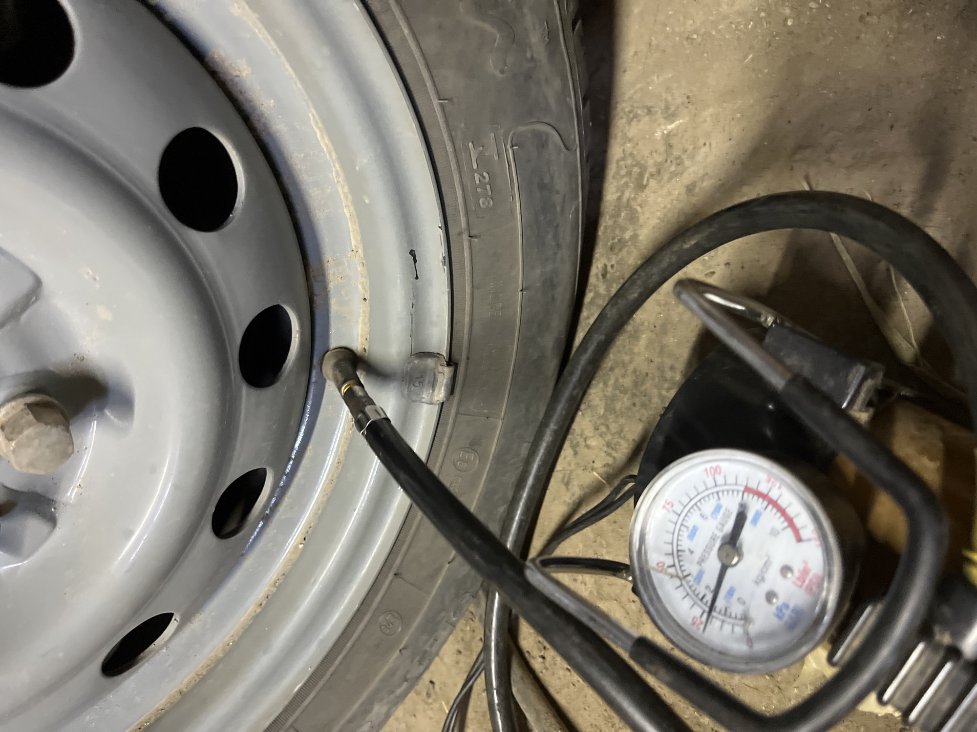 This media file shows a close-up view of a car tire being inflated. The scene is set indoors, likely in a garage or workshop, as indicated by the concrete floor.
The main subjects are a car tire and a tire pressure gauge connected to an air hose. The tire is gray on the rim and black on the tread. The pressure gauge has a white face with black markings and a red needle pointing to approximately 2.5 kg/cm², which is roughly 35 PSI. The air hose is black and appears to be connected to the tire's valve stem.
The activity taking place is tire inflation, with the gauge monitoring the pressure. The focus is on the technical process of maintaining proper tire pressure.
Notable details include markings on the tire sidewall, one of which appears to be "278." There are also circular ECE approval marks visible. The lighting is artificial, suggesting an indoor environment. No people are visible in the frame, and there are no cues about the time of day or weather. The overall mood is neutral and functional, focusing on the task at hand. The location context provided is Krasnyye Barrikady, Russia, but this information is not visually discernible from the image itself.