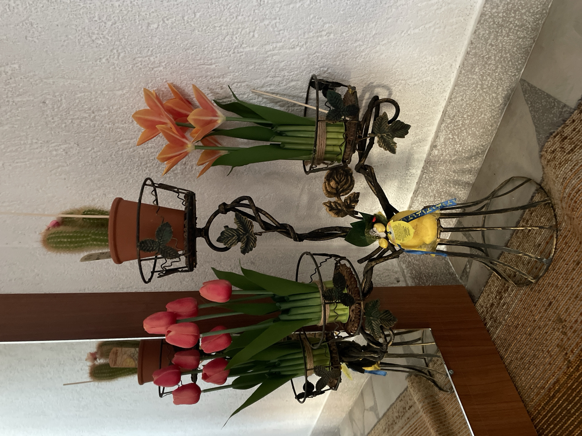A wrought iron candelabra with multiple tiers holds several artificial flower arrangements and a potted cactus. The candelabra's base is intricately designed with decorative leaves and stands on a woven rug. To the left of the candelabra is a tall wooden-framed mirror reflecting a portion of the scene, including the potted cactus and more artificial tulips. To the right of the candelabra, a textured white wall serves as the background. The overall setting appears to be an indoor decorative display, possibly in a home or establishment in Zlatograd, Bulgaria. There are no people present in the image.