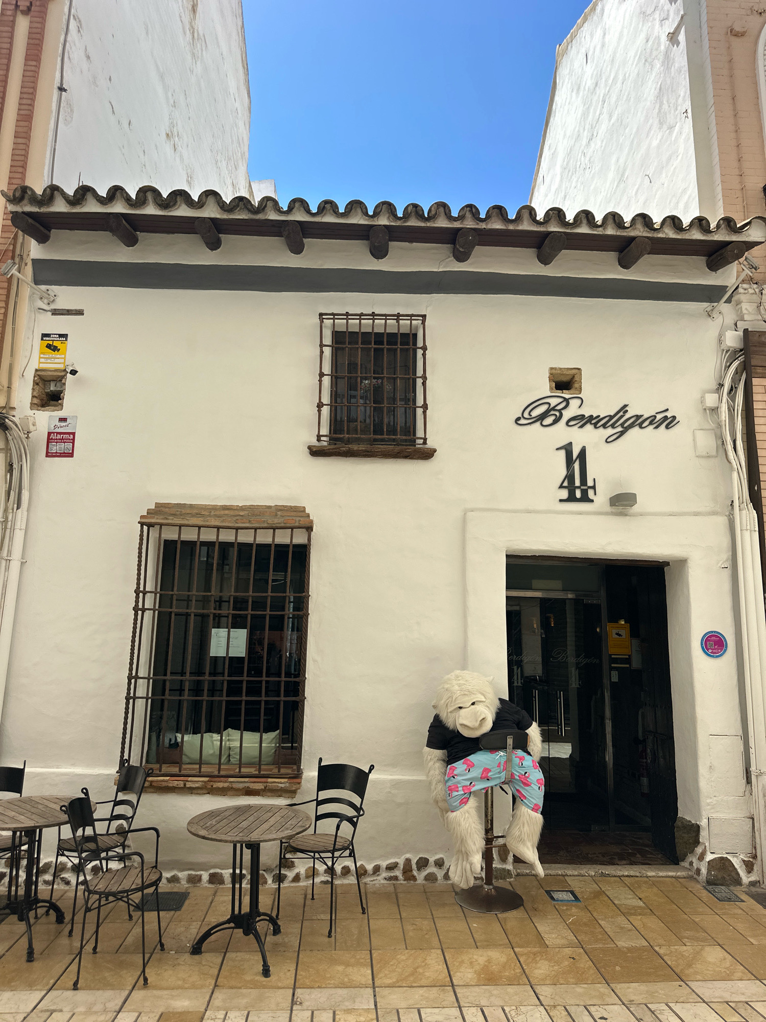 A white facade building with a tiled roof and exposed wooden beams stands in a street. A large white stuffed ape wearing a black shirt and pink flamingo-patterned shorts sits on a bar stool in front of the entrance. The ape is positioned on a tiled pavement. Several tables and chairs are set up outside the building, suggesting an outdoor seating area for a business. The building has barred windows, one of which has green cushions visible inside. Signs are affixed to the wall, including one with a yellow background and a black camera icon, indicating a security camera is present, and another red sign labeled "ALARMA". A black script sign above the entrance reads "Berdigón", and below it, the number "14" is displayed in black. The street is paved with light-colored tiles. The sky above is clear and blue. The building is likely located in the city of Huelva, Spain, given the architectural style and the language on the signs.