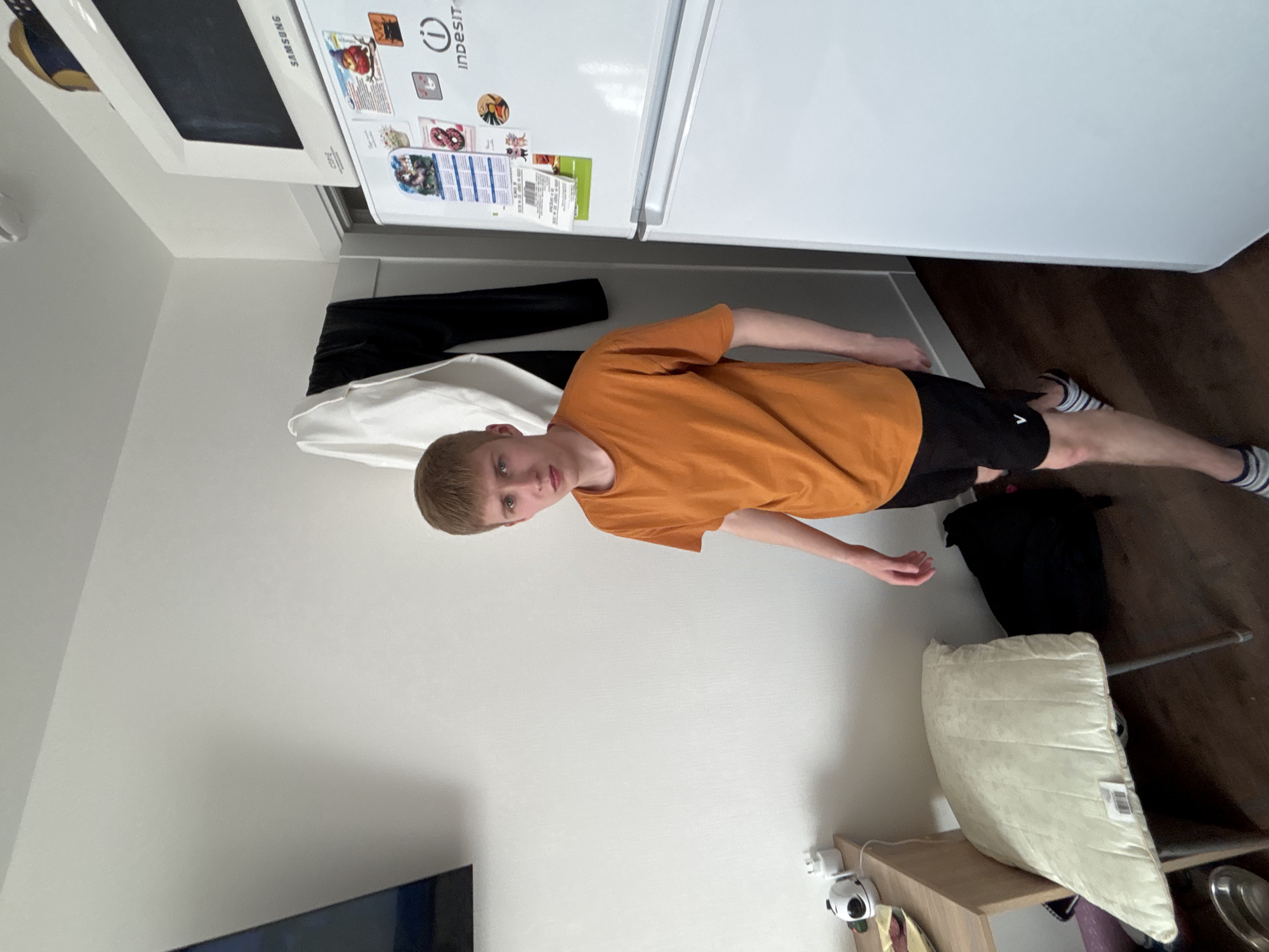 A boy stands in an indoor location. He wears an orange t-shirt, black shorts, and striped socks. He is positioned near a refrigerator displaying magnets, a white top and black pants are on the side. The environment includes wood flooring and a shelving unit with a white pillow on top. A security camera is attached to the wall. The location is not identifiable as Vsevolozhsk, Russia from the visual information.