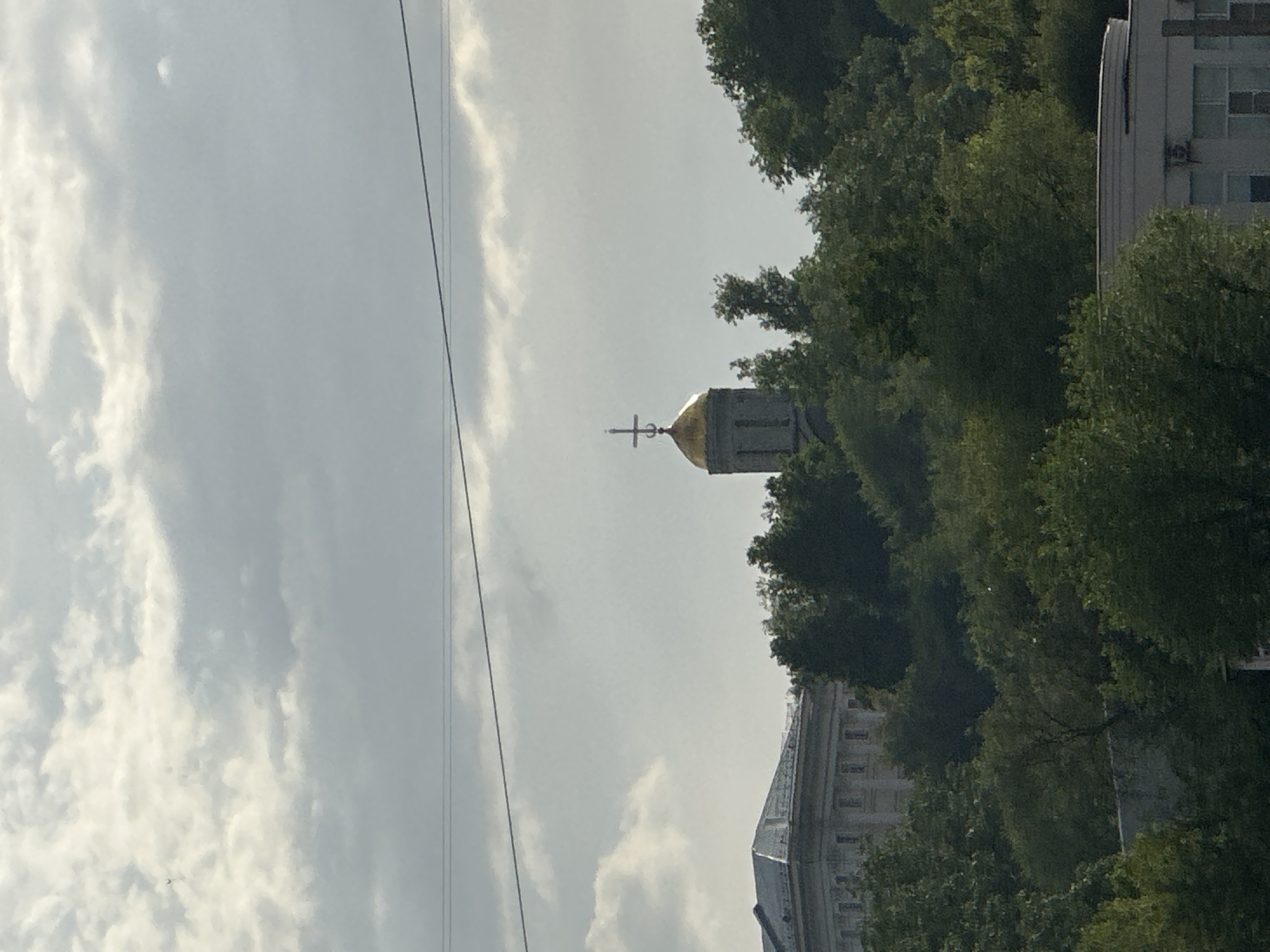 The image shows a partly cloudy sky above a cityscape in Vladimir, Russia. A golden-domed Orthodox church with a cross atop is visible, partially obscured by trees. In the lower right corner of the image, part of a multi-story building with windows can be seen. Power lines run across the sky.