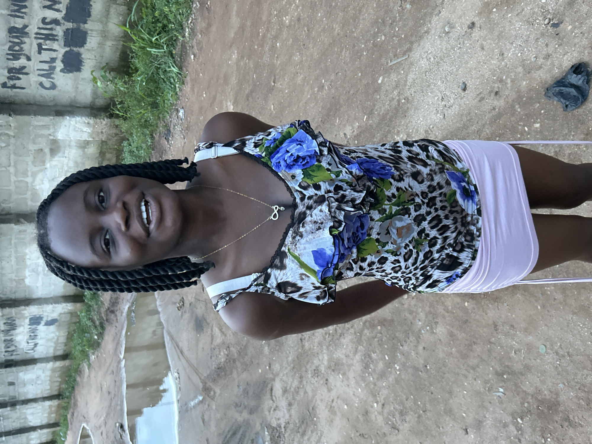The image, taken in Ihiagwa, Nigeria, features a young Black woman standing outdoors, smiling broadly at the camera. She has her hair styled in black braids. She is wearing a sleeveless top with a distinctive leopard print pattern adorned with prominent blue and green floral designs. A white bra or camisole strap is visible under her top on her right shoulder. Around her neck, she wears a delicate gold chain with a small pendant that resembles the number '8' or an infinity symbol. Her bottom attire consists of a light pink or lavender-colored skirt.
The setting appears to be an outdoor, somewhat undeveloped area. The ground beneath her is uneven dirt, and there is a noticeable puddle of water to her left, reflecting the sky. Behind her, a weathered concrete block wall stretches across the background. Some green vegetation, possibly grass or weeds, grows along the base of this wall. On the wall, there is visible graffiti in black lettering. Partially obscured, the text reads "FOR YOUR IN" on the top line, and "CALL THIS N" on the line below, with what appear to be blacked-out sections where phone numbers or other details might have been. The lighting suggests it is daytime, and the overall mood conveyed by the woman's smile is cheerful.