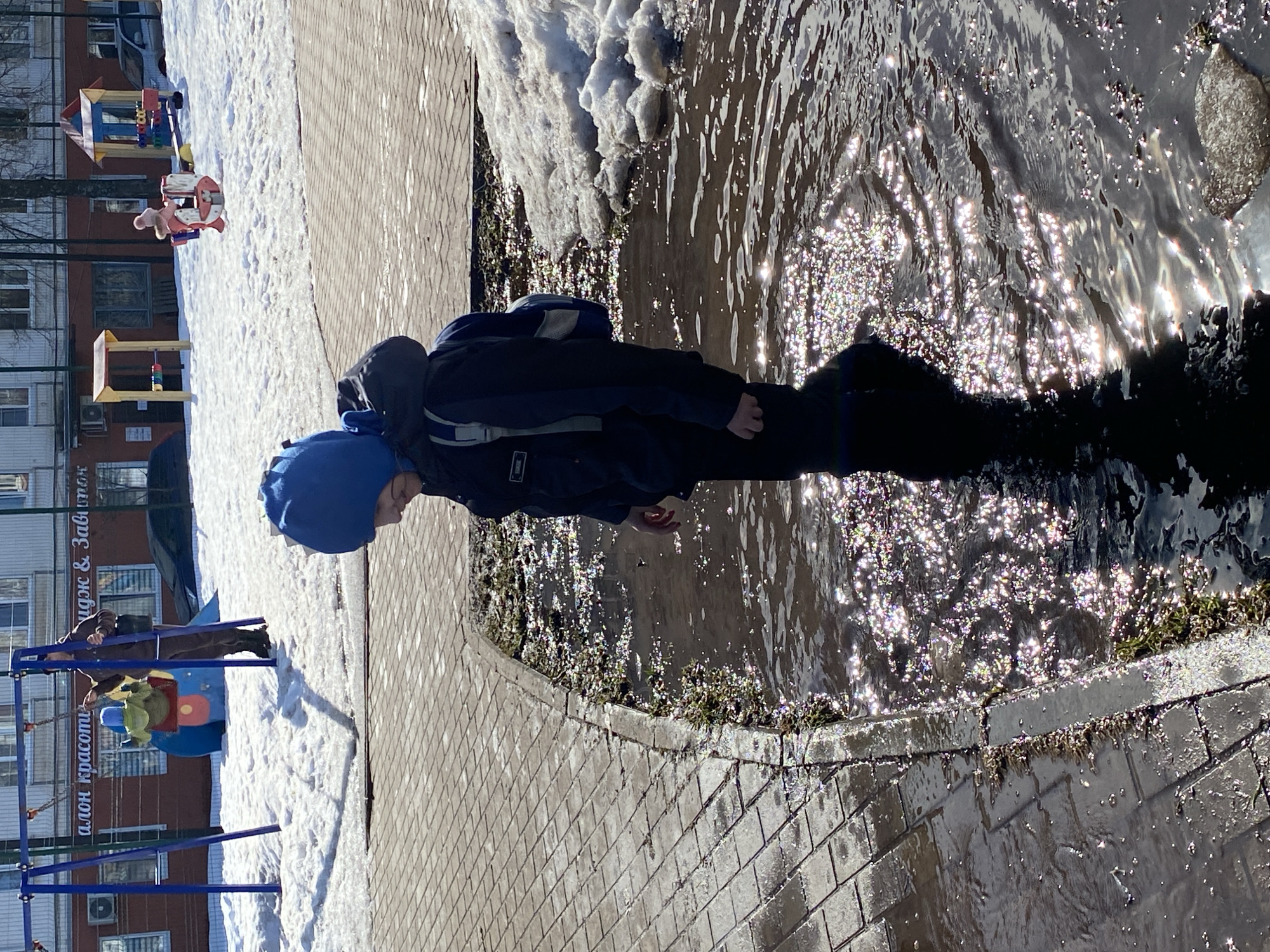 The scene depicts an outdoor urban environment, specifically a playground area in the city of Chaikovsky, Russia, characterized by melting snow and puddles under clear daylight conditions.
In the immediate foreground, a child, viewed from the right side in profile, stands immersed in a large, sunlit puddle. The child wears a dark blue hooded jacket, dark trousers, and a blue hood, with a grey and blue backpack on their back. Their gaze is directed slightly downwards or to the right, exhibiting a faint smile. Sunlight creates numerous bright, shimmering reflections on the dark water's surface. The puddle borders a grey paved pathway constructed from rectangular pavers. Small bits of vegetation are visible along the puddle's edge.
Beyond the paved path, the ground is covered in patches of melting snow. In the midground, a blue metal swing set with two swings is present. An adult, dressed in dark clothing, stands behind a child in a green outfit on one of the swings, appearing to push them. To the right of the swing set, other colorful playground structures are visible, including a red and white spring rider with a child in a pink hat seated on it, and a small, multi-colored seesaw or similar apparatus.
The background features a multi-story building with a light-colored upper facade and an orange-brown lower level. A sign on the building's ground floor reads "салон красоты" (beauty salon) and partially "идж & Завиток" in Russian. Several vehicles, including a dark sedan and a silver car, are parked near the building. Bare trees are visible in the distance.