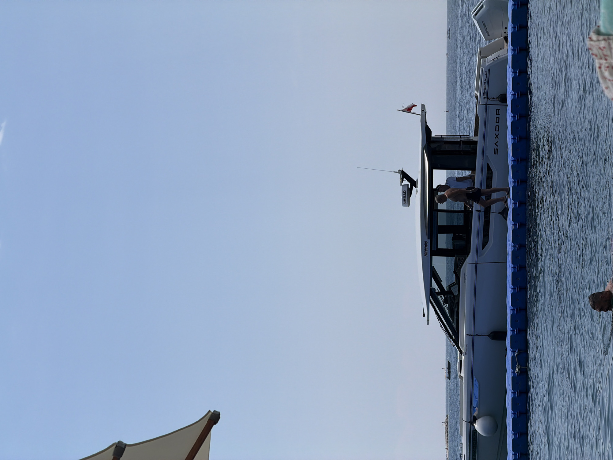 The image shows a modern yacht docked in clear blue water, likely in Budva, Montenegro. Two individuals are visible on the deck of the yacht, with one standing and another seated. The yacht itself is sleek and white, with tinted windows and a name, "SAXDOR," visible along its hull. A red and white flag flies from the top of the yacht. A radar unit labeled "SIMRAD" is also mounted on the vessel. The yacht is connected to a blue floating dock. The water is calm, reflecting the clear, bright sky, suggesting a pleasant day, possibly late morning or early afternoon. In the background, the coastline and other boats can be seen in the distance, typical of a marina or coastal town. Another person is partially visible in the water on the right side of the frame. The overall scene conveys a sense of leisure and a bright, sunny day at sea.