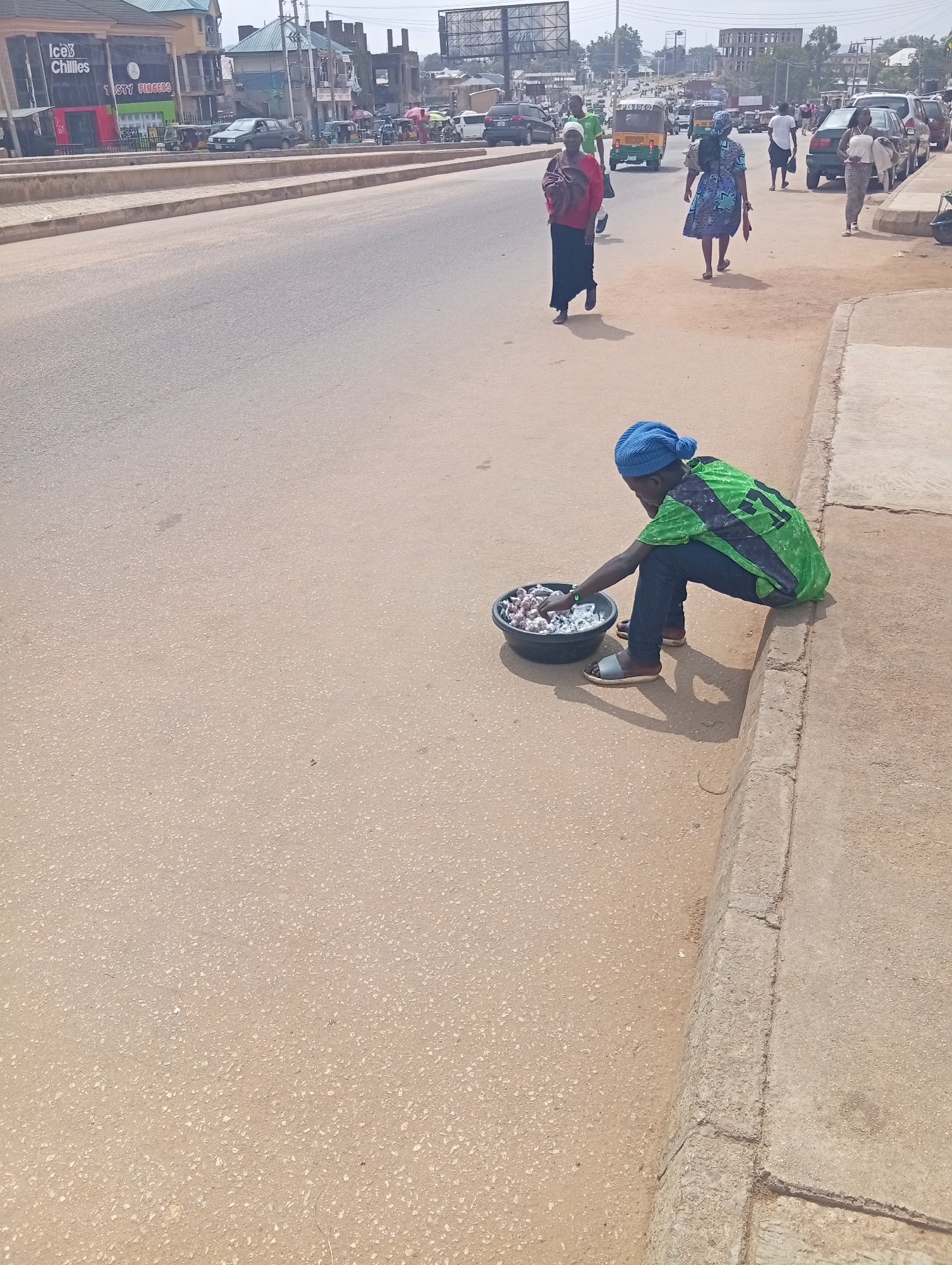 The image captures a street scene in Kabong, Nigeria, on a bright, sunny day. A young individual wearing a blue knit cap and a green and black patterned shirt is crouched at the edge of a concrete sidewalk, facing a black bowl filled with small, white objects. The individual appears to be a vendor, possibly selling the items in the bowl, which resemble small candies or snacks. They are wearing dark jeans and grey slide sandals. The sidewalk itself is worn and cracked in places.
In the background, the street is bustling with activity. Several vehicles, including a green car, a white van, and at least two auto-rickshaws (one yellow and one white), are visible on the asphalt road. Pedestrians are also present, with a woman in a blue and white patterned dress walking away from the camera, and another woman in a red top and black skirt walking towards it. Further down the street, more people and vehicles are present, along with buildings that appear to be shops and residences. The overall environment suggests a commercial or residential area within the city, with active daily life.
