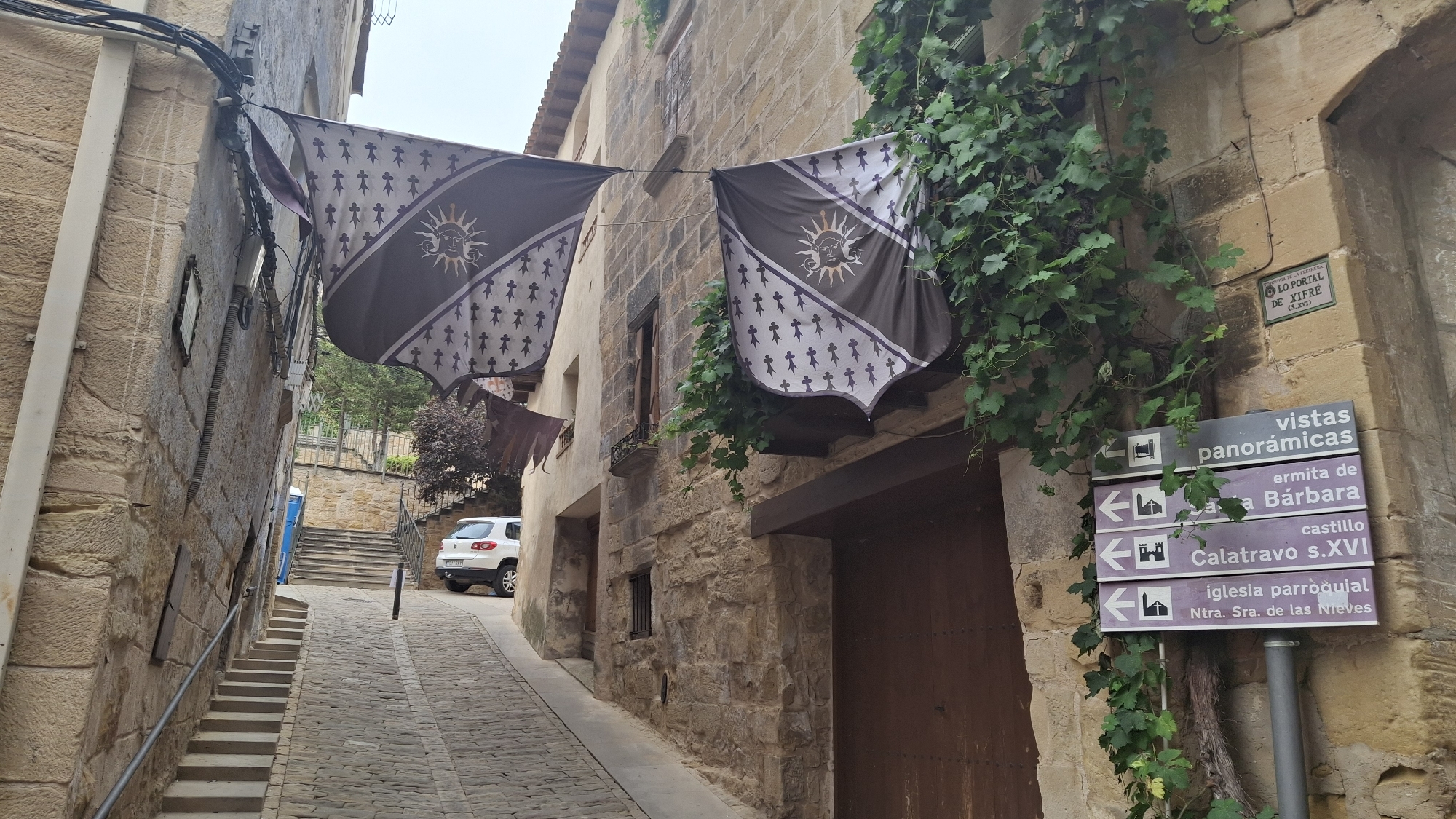 The image depicts a narrow, stone-paved street in La Fresneda, Spain, winding uphill between two ancient stone buildings. Two large, dark fabric banners with a sun emblem and a pattern of small figures hang across the street, suspended from wires attached to the buildings. The banner on the left is partially obscured by the building wall and a cluster of dark-leaved bushes. The banner on the right is more visible, adorned with a sun with a face and an intricate border. Lush green vines cascade down the wall of the right building, partially covering a weathered stone facade and a wooden door. A brown signpost with white lettering lists destinations: "vistas panorámicas", "ermita de Santa Bárbara", "castillo Calatravo s.XVI", and "iglesia parroquial Ntra. Sra. de las Nieves", with directional arrows indicating left turns for most. A white SUV is parked further up the street on the right, near a set of stone stairs leading upwards. A blue portable toilet is visible on the left side of the street, near a set of concrete steps. The sky is overcast, and the overall impression is one of historical architecture and a tranquil, secluded urban setting.