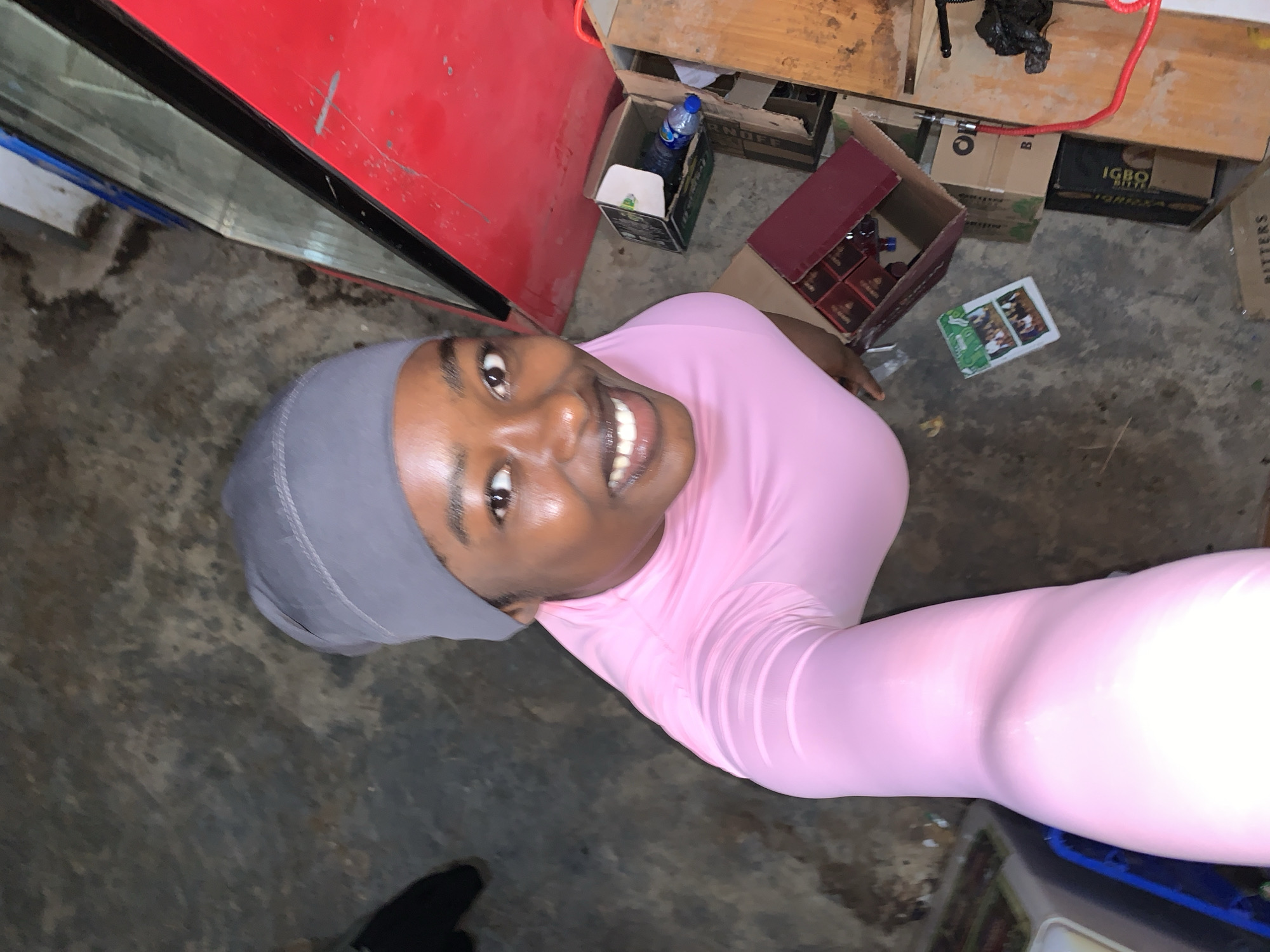 A young woman with brown skin and a gray headscarf is smiling at the camera. She is wearing a tight-fitting pink long-sleeved top. The photo appears to be taken from a high angle, possibly a selfie. The location is cluttered, with cardboard boxes, bottles, and other items scattered around on a dirty concrete floor. Some boxes are labeled "IGBO BITTERS." A red refrigerator is visible in the upper left corner. A small part of what seems to be a black shoe is visible in the lower left corner. The image suggests an indoor setting that is potentially in a commercial or storage area. Given the presence of Igbo Bitters, this area is very likely in or near New Stadium, Nigeria.