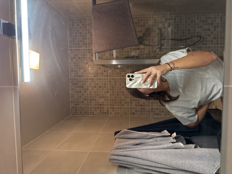 An image captured as a mirror selfie depicts a female individual in a tiled bathroom, located in Cannes, France. The subject, viewed from the mid-chest up, holds an iridescent smartphone with her left hand, obstructing her face. Her right hand is positioned on her hip. She wears a light-colored, possibly striped, short-sleeved top. Visible accessories include gold hoop earrings, a ring on her left ring finger, white nail polish, and a thin bracelet on her left wrist. The bathroom features two distinct tile patterns: larger, light beige rectangular tiles on the upper wall, and smaller, brown and beige mosaic tiles on the lower wall and inside the shower enclosure. To the left, multiple grey and one dark blue towel hang on a hook or rack. On the right, a clear glass shower enclosure with a chrome frame houses a dark brown towel draped over a metal rail, along with a chrome shower head and hose. A horizontal LED light fixture is mounted above the mirror, which exhibits visible smudges and reflections of light sources.