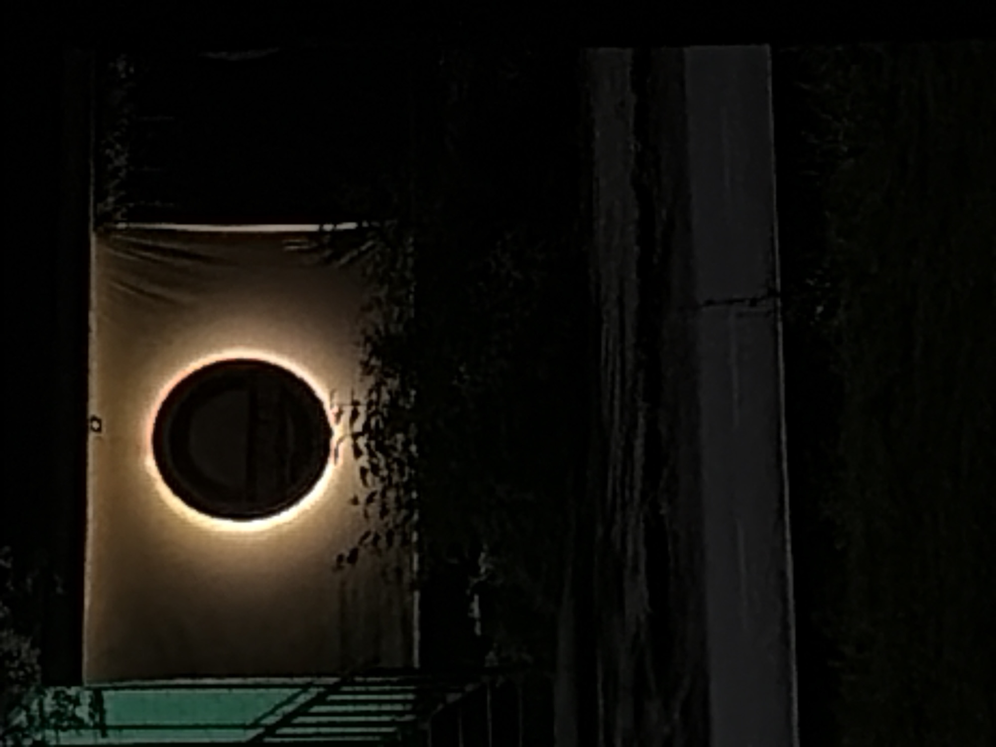 At night, a lit circular window is centered on the front of a building. The building is a light color. In front of the building is some greenery and what appears to be a fence. To the right of the building is a dark pillar. Further to the right is more greenery. The location is unknown.