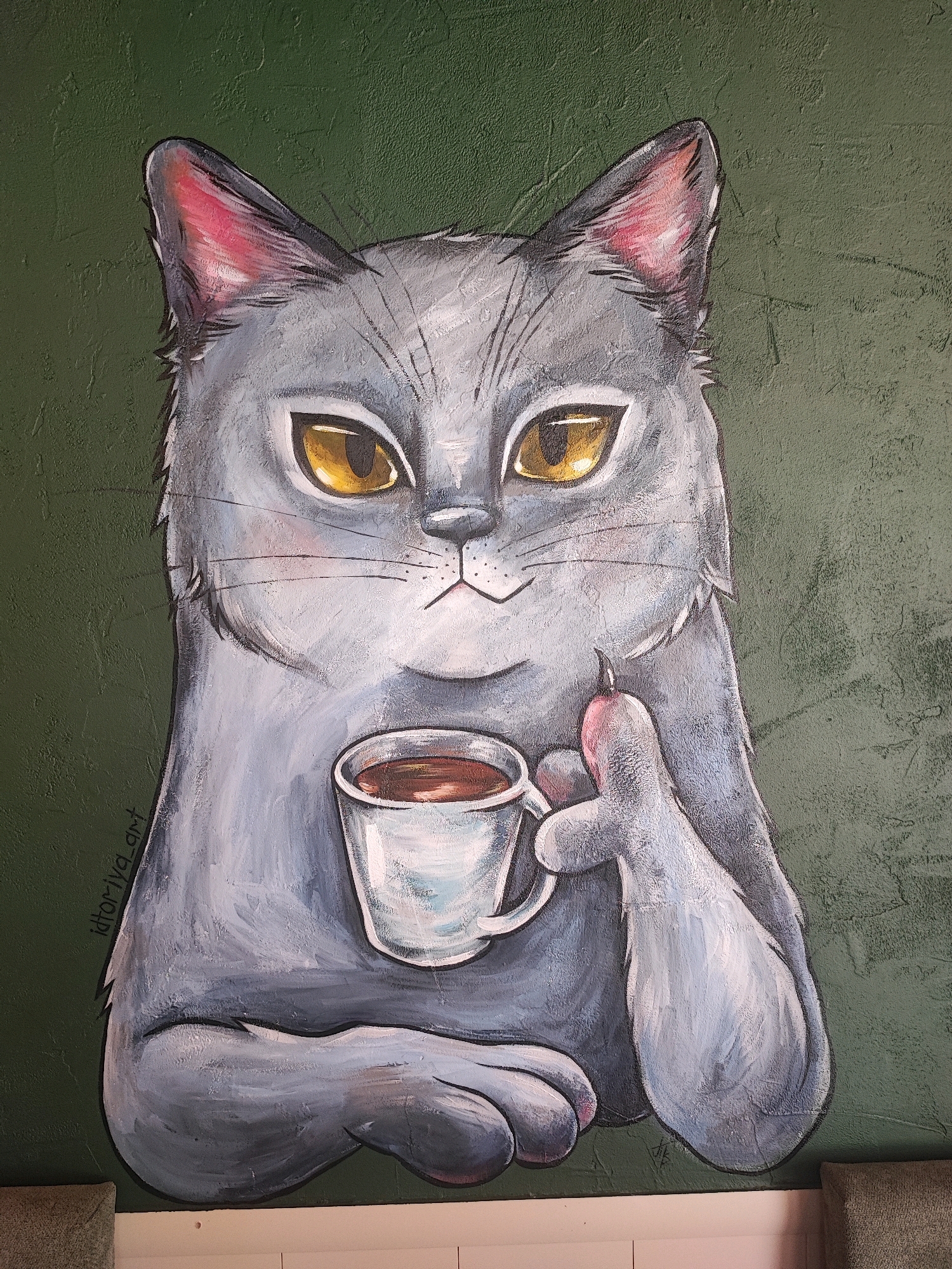 A painted mural of a gray cat with yellow eyes sits against a textured dark green background. The cat is holding a white mug of brown liquid in its left paw and a small pink object, possibly a treat, in its right paw. The cat's ears are outlined in black with pink interiors. The mural is located on the exterior wall of a building in Pyatigorsk, Russia. A signature, "Idtoriya_art", is visible in the lower left portion of the mural. The lower edge of the image shows a portion of a pink surface and a textured beige object.