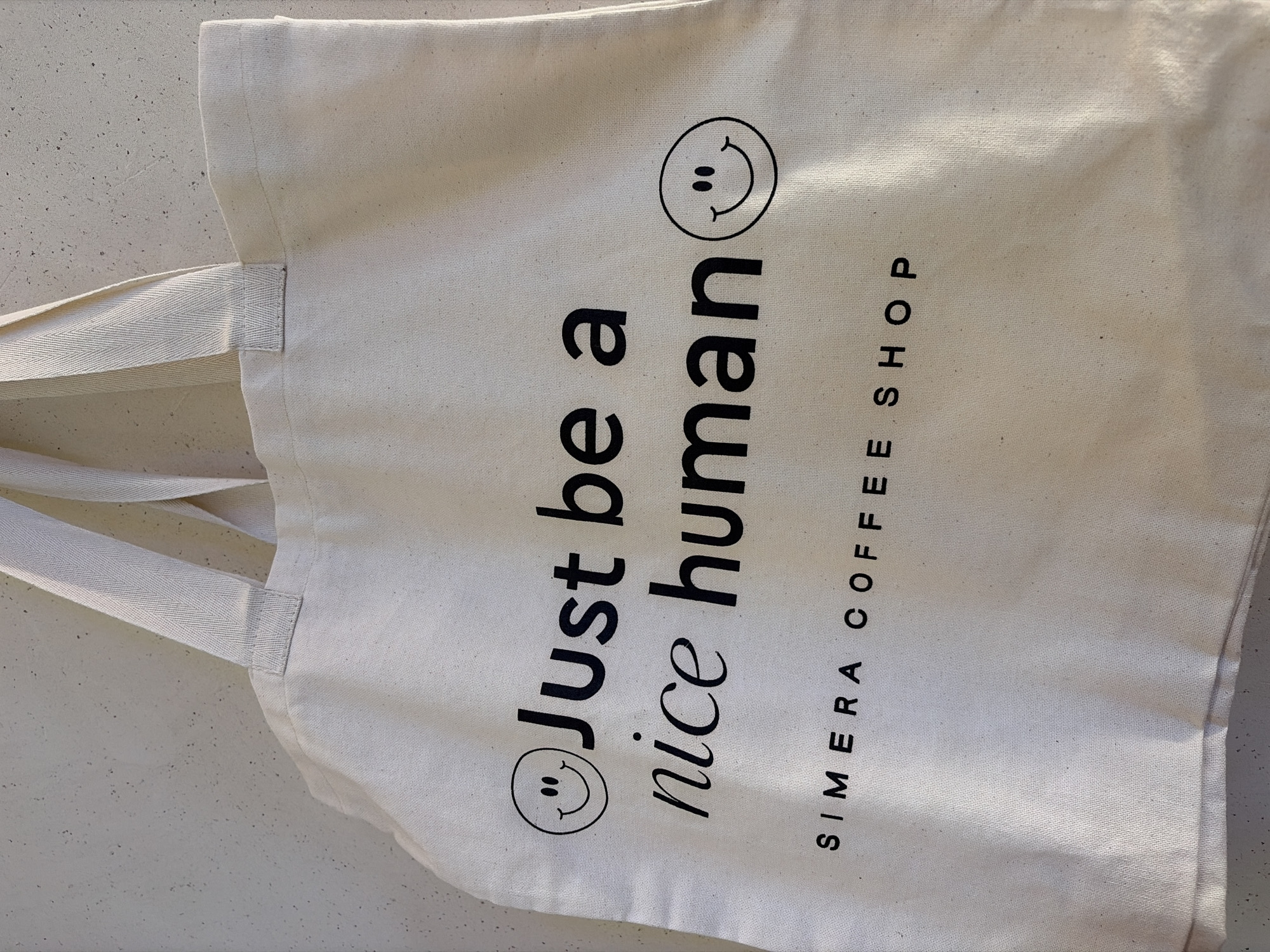 The image shows a light beige canvas tote bag laying on a light gray surface. The bag has two handles of the same material. Printed on the bag in black lettering is the phrase "Just be a nice human," with a smiley face at the beginning and end of the phrase. Underneath, in smaller black text, is "SIMERA COFFEE SHOP." The location is unknown.