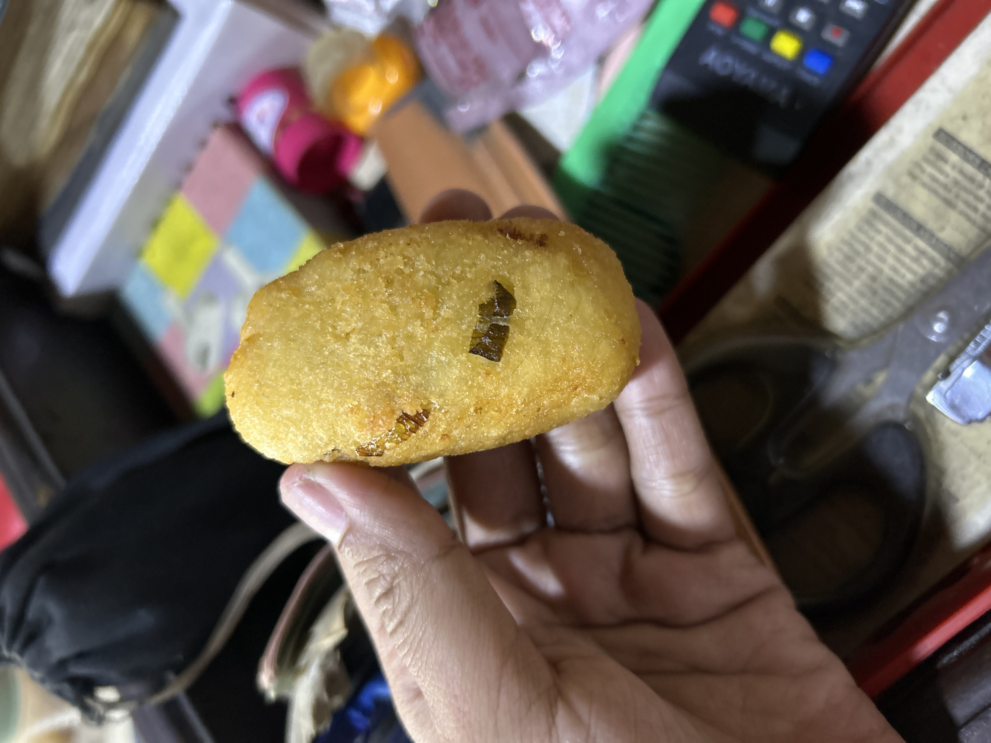 A person's hand holds a golden-brown fried snack. The snack is oval-shaped and appears to be made of dough, with some darker specks of seasoning or ingredient visible on its surface. The hand is positioned in the foreground, with fingers curled around the snack. In the background, various out-of-focus objects are visible, including a black drawstring bag, a box with colorful squares, a pink object, and a black remote control. The overall setting appears to be indoors, possibly a home or a shop, and the lighting suggests it might be nighttime or an artificially lit environment. The specific location in Depok, Indonesia cannot be determined from the visual information provided.