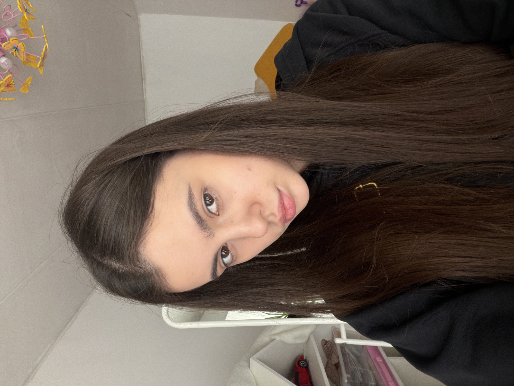 A young woman with long, dark brown hair is positioned in the center of the frame, facing the camera with a slight tilt of her head. She has fair skin, brown eyes, and full lips. She is wearing a black hooded sweatshirt. In the background, there's a white wall with a decorative item featuring pink and yellow butterflies. Below, a white shelf unit houses various objects including a red toy car and assorted items in containers. The location is implied to be in Chaikovsky, Russia, based on the prompt's instructions.