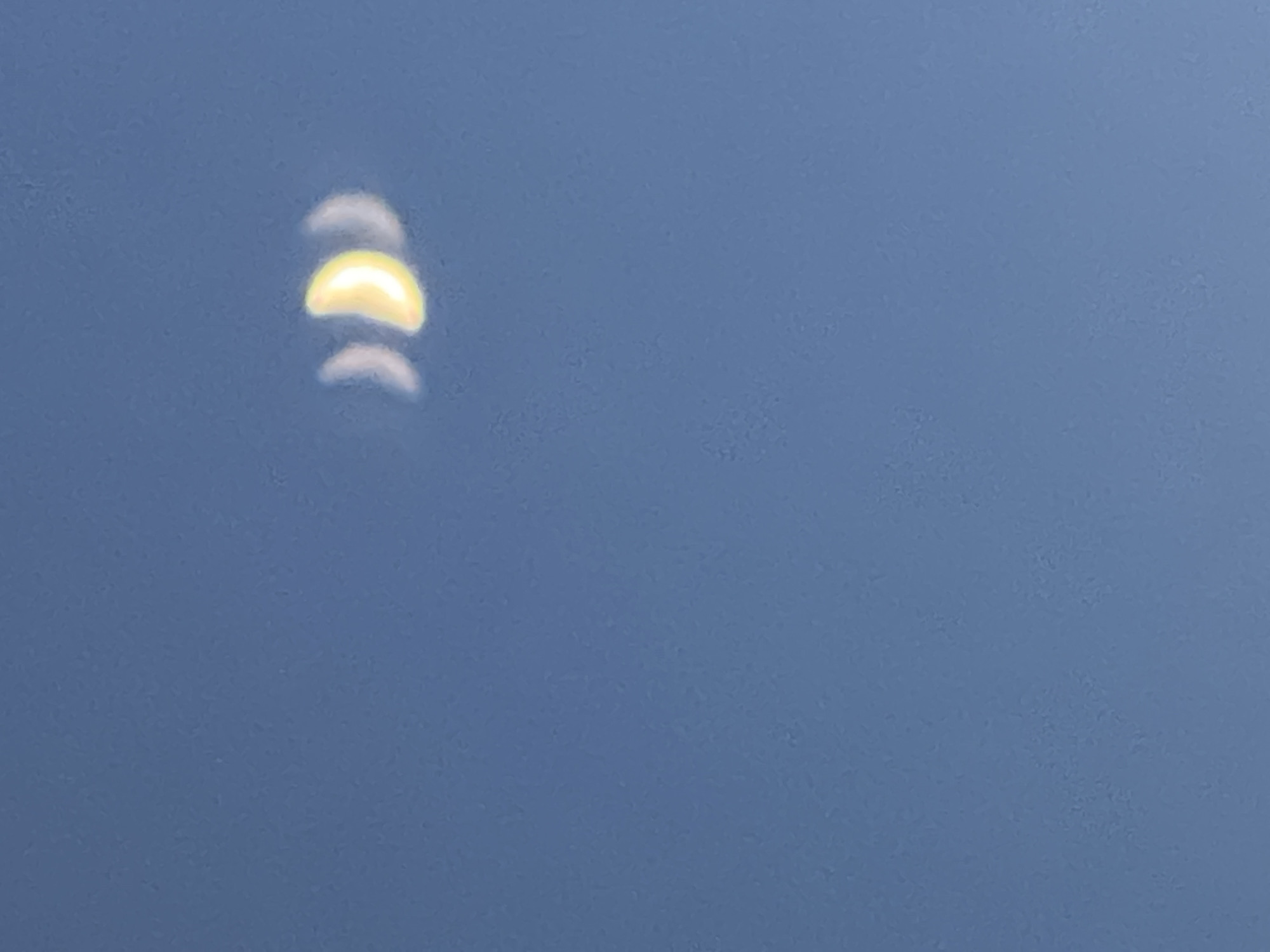 This is a photograph of the sky with a blurred crescent shape, likely the moon, visible in the upper right quadrant. The sky is a clear, bright blue. There are no discernible landmarks or elements that would allow for specific identification of the location within Minsk, Belarus. The image is out of focus, rendering details of the crescent shape indistinct. The overall impression is of a bright, open sky with a celestial object.