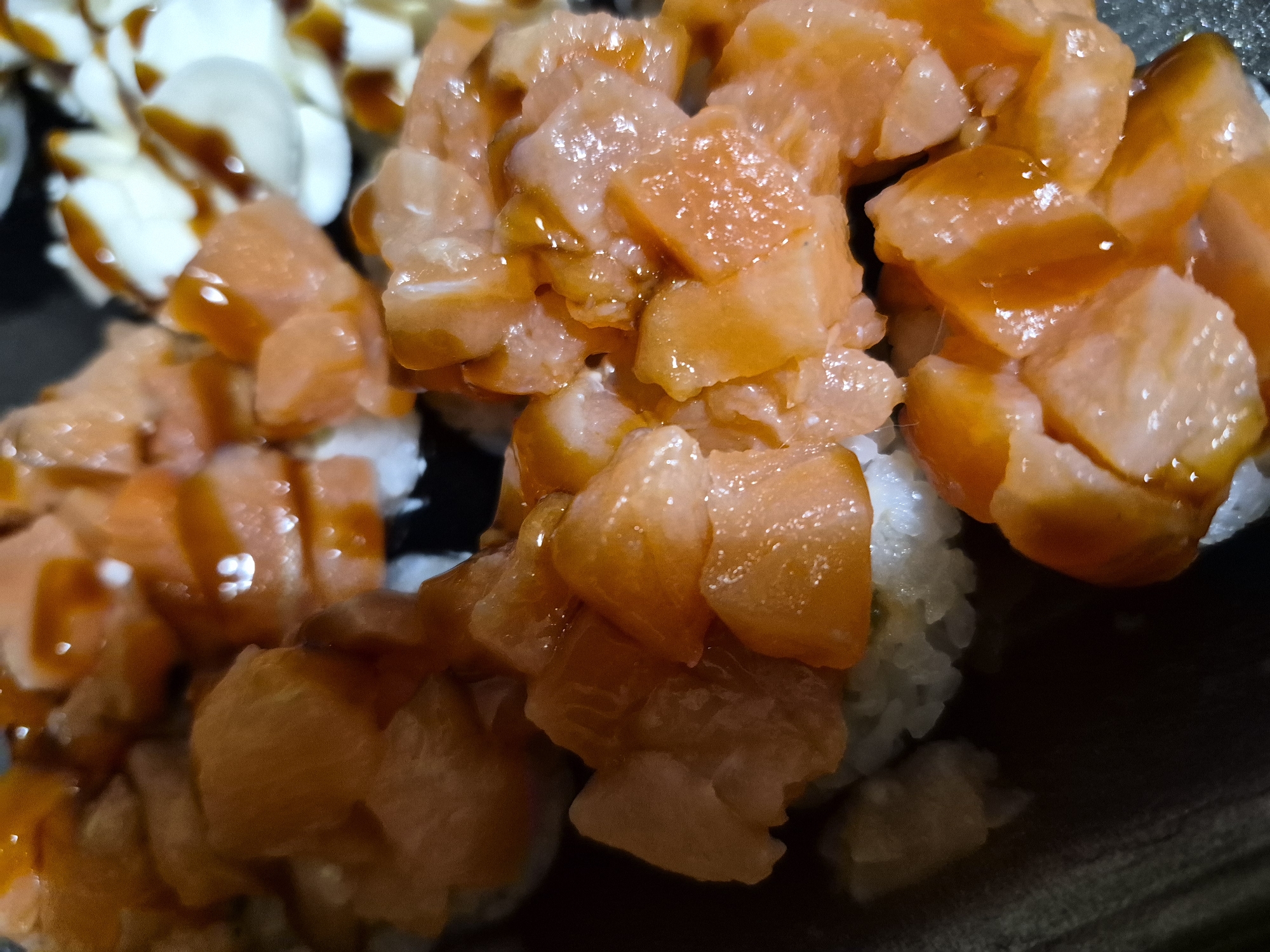 A close-up shot features a sushi dish, likely salmon sushi, with rice and a brown sauce drizzled on top. There is also sushi with white topping, likely mozzarella, drizzled with the same brown sauce. There is no evidence that the picture was taken in Tobolsk, Russia. There are no people in the image. The environment is out of focus.