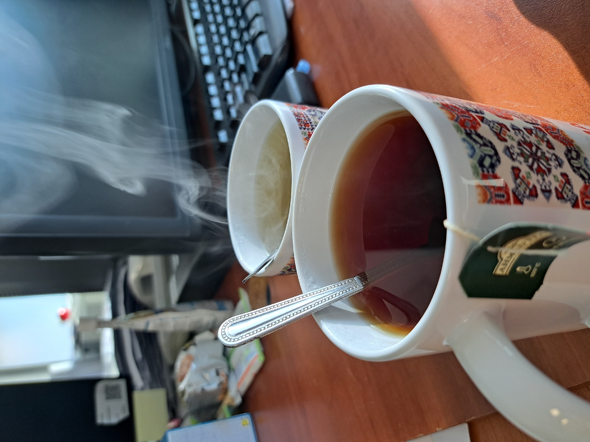 Two mugs are positioned on a wooden desk. The mug on the right is filled with dark liquid, likely tea, and contains a spoon and a tea bag. The mug on the left appears to contain a lighter-colored liquid. Steam is rising from the left mug. In the background, a computer monitor and keyboard are visible. The location is Unknown, Unknown.
