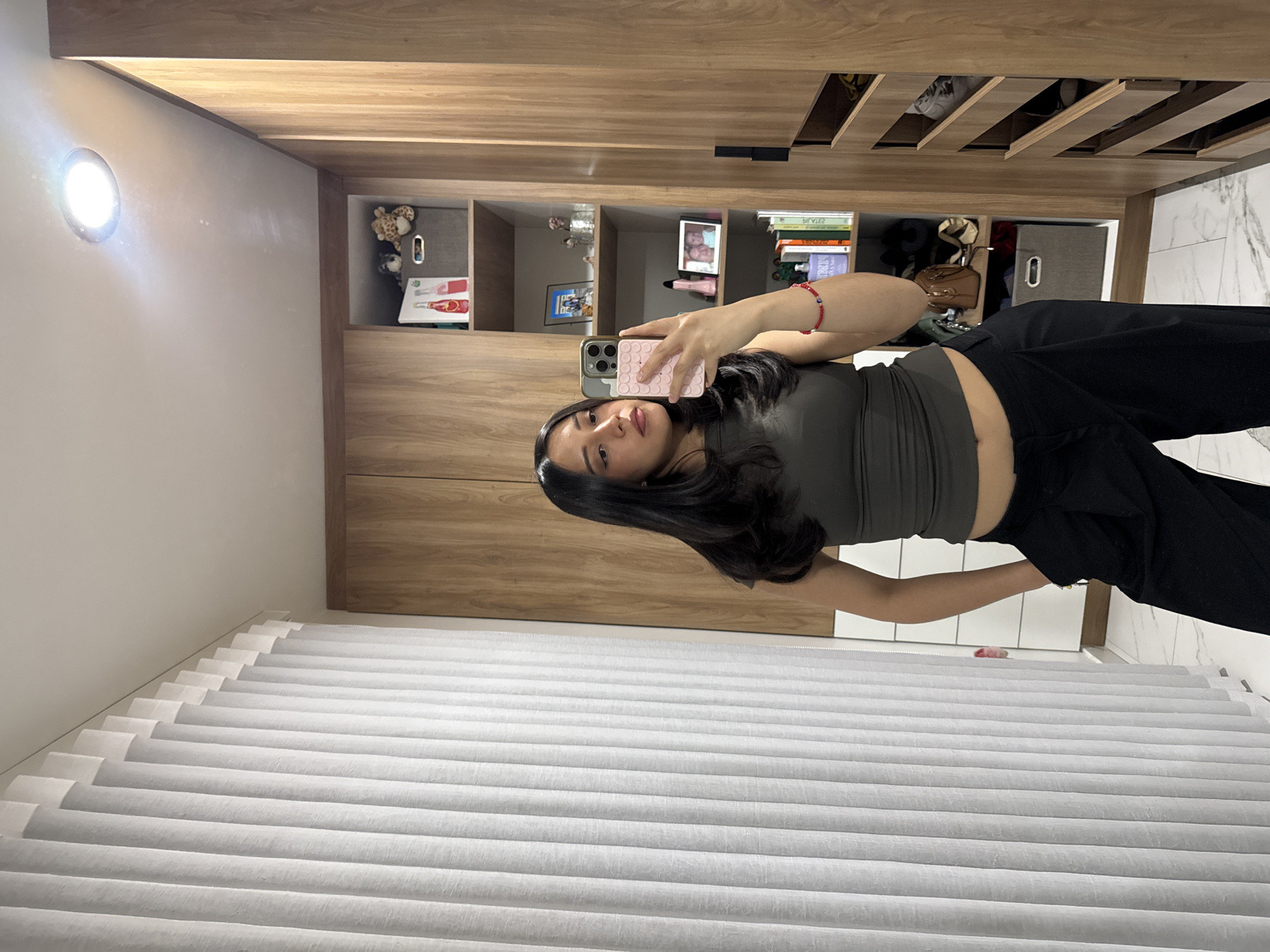 A female individual is depicted taking a mirror selfie within a private residence in the city of Puebla, Mexico. She is positioned centrally, facing forward, holding a light pink smartphone with a patterned case in her right hand. A red beaded bracelet with a small blue "evil eye" bead is visible on her right wrist. Her left hand is inserted into the pocket of her black wide-leg pants. She wears a dark olive/grey sleeveless crop top, exposing her midriff. Her dark hair is styled down. The background on the right and center features a light wood wardrobe system with open shelving and closed compartments. The open shelves contain various personal items including stuffed animals, red-labeled bottles, a grey fabric storage bin, framed photographs, a pink cylindrical object, books, and a brown handbag. A section of the wardrobe to the far right features multiple horizontal open shelves displaying footwear. To the left, a large window is covered by vertical white blinds. The floor is composed of white tiles with grey veining. A single bright, recessed circular light fixture is visible on the white ceiling.