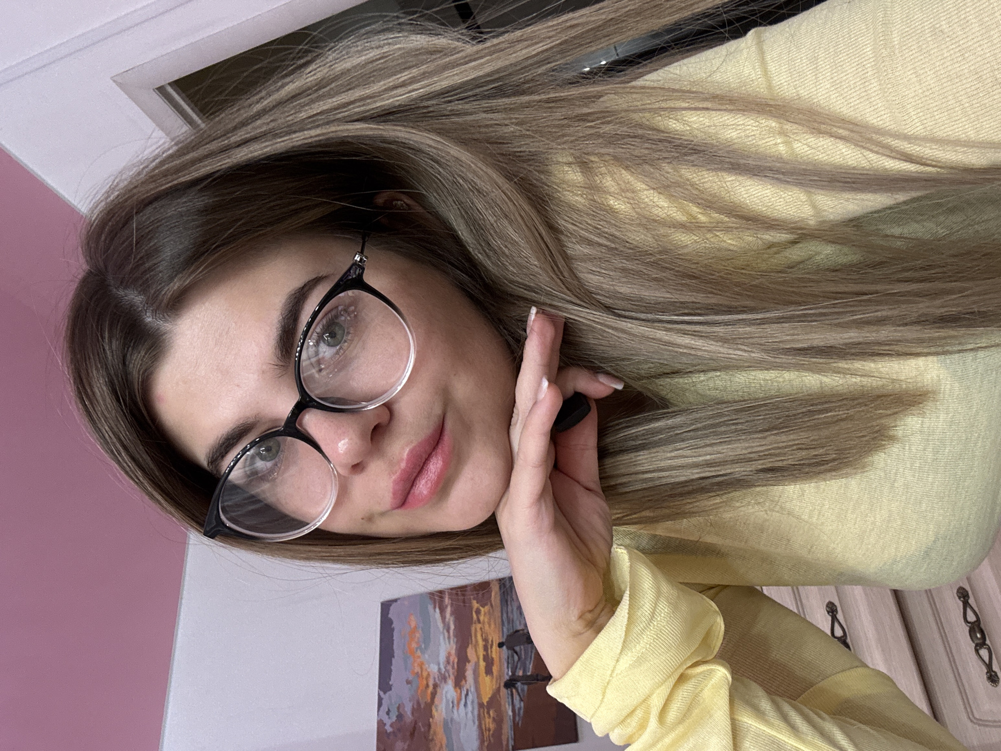 An image taken in Chaikovsky, Russia, depicts a female individual from the chest up, positioned slightly off-center and looking directly at the camera. She has long, straight, light brown hair with blonde highlights, parted down the middle. She is wearing clear-framed eyeglasses with black upper rims and a yellow long-sleeved top. Her right hand is raised, with fingers curled, supporting her chin. Her fingernails are painted white. The background features a two-toned wall, with the upper section painted pink and the lower section light grey or white. A rectangular painting of a sunset beach scene with two silhouetted figures is mounted on the lower wall section behind her left shoulder. In the lower left, a light-colored dresser with two ornate dark-colored handles is partially visible. A white door frame is present in the upper right background.