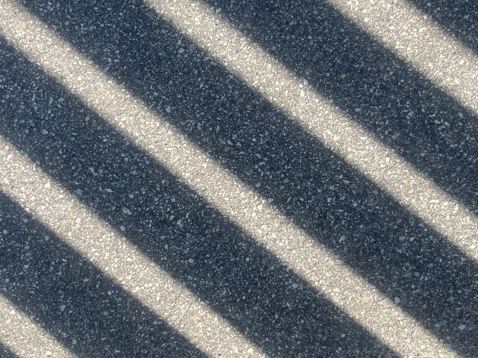 This is an overhead shot of asphalt with diagonal shadows cast across it, possibly from a fence or blinds. The asphalt is dark gray with small white and light gray aggregate visible. The shadows are a lighter gray, indicating they are not fully opaque. There are no people or discernible objects within the frame. The scene appears to be outdoors in a city, but the specific location in Kyiv, Ukraine cannot be determined from the image alone.