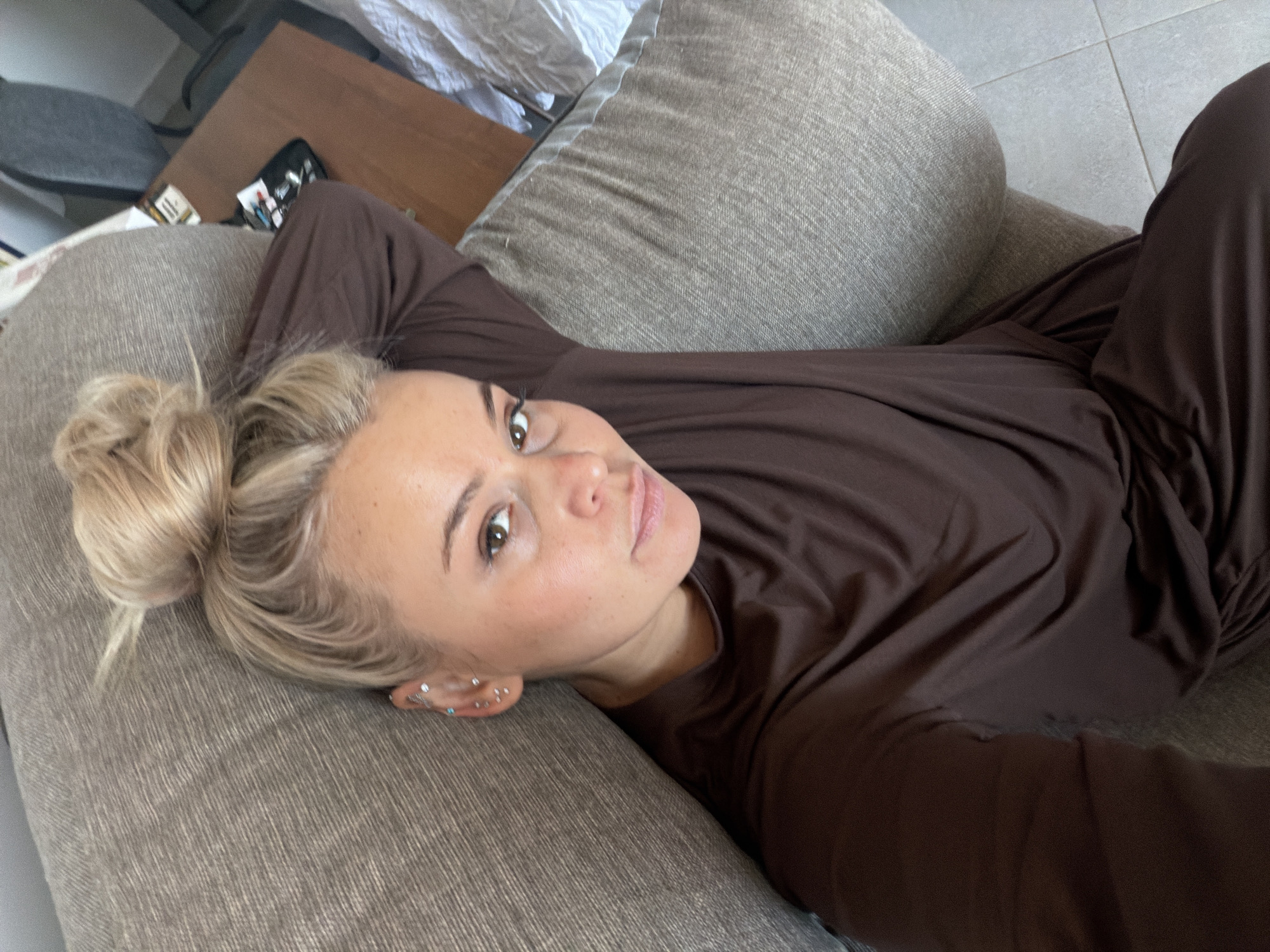 A woman with blonde hair, tied in a messy bun, is lying on her back on a light brown, textured fabric couch. She is wearing a comfortable, plain brown long-sleeved top and matching trousers. Her gaze is directed towards the camera, with a neutral and relaxed expression. Her left ear, visible in the foreground, features multiple small stud piercings and a more prominent earring with blue accents.
The setting appears to be an indoor space, likely a living room or bedroom. In the upper left background, a wooden table is visible, holding a small black zippered case from which pens or similar items protrude. Next to it, a white rectangular object, possibly a pack of cigarettes or a small box, is present. A dark grey chair is partially seen behind the table, and a crumpled white sheet or blanket is in the top left corner. To the right, a grey tiled floor is visible. The lighting suggests it is daytime. The overall scene depicts a casual moment of relaxation at home. No specific visual cues indicate the location of Sarapul, Russia, which is the given context. There is no visible text in the image.