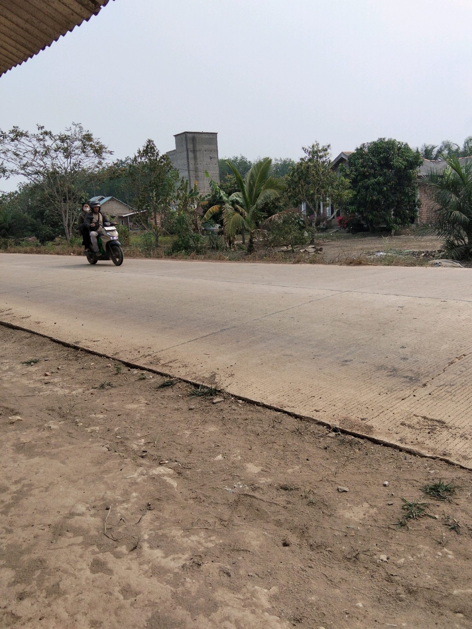 In Perambahan, Indonesia, a motorcycle with two individuals is traveling on a paved road. The driver and passenger are both wearing helmets and masks. The motorcycle is green and white. The road is a light gray concrete surface with visible expansion joints and grooves. To the left of the road is a dirt path with scattered dry vegetation and small stones. Behind the road, on the left side of the frame, there are trees and a tall, square, gray building that appears to be a silo or industrial structure. Further back, to the right, are houses with visible rooftops and lush green trees, including palm trees. The sky is overcast and pale. The scene depicts a rural or semi-rural area in Indonesia during daylight.