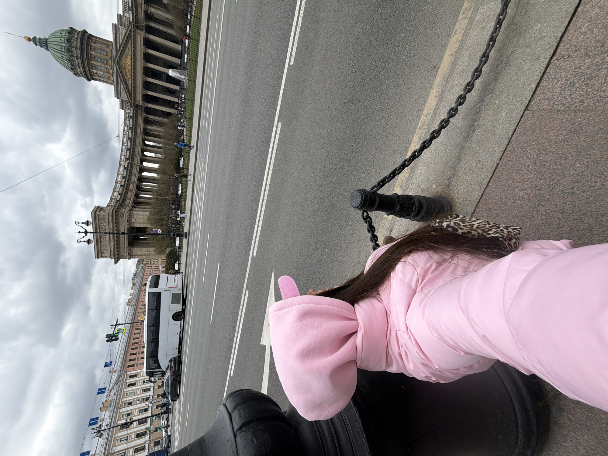 An upright, eye-level shot captures a street scene in Saint Petersburg, Russia, on an overcast day. In the foreground, a person is seen from behind and to the side, wearing a vibrant pink puffer jacket with a hood featuring distinctive rabbit ears. Their long, dark hair is visible, and a leopard-print bag rests near their hip. They are positioned next to a black metal chain barrier, which separates the sidewalk from a wide asphalt road.
The road features clear white lane markings. Across the road, the magnificent Kazan Cathedral dominates the background, identifiable by its grand neoclassical architecture, featuring a sweeping colonnade, a pediment, and a prominent green dome topped with a golden spire. Other historic buildings, characteristic of Saint Petersburg, line the street further into the distance. A large white double-decker tour bus is visible alongside other vehicles on the street. Traffic lights and various street signs are also present. The sky is heavily clouded and grey, suggesting cool weather. No discernible text is visible.