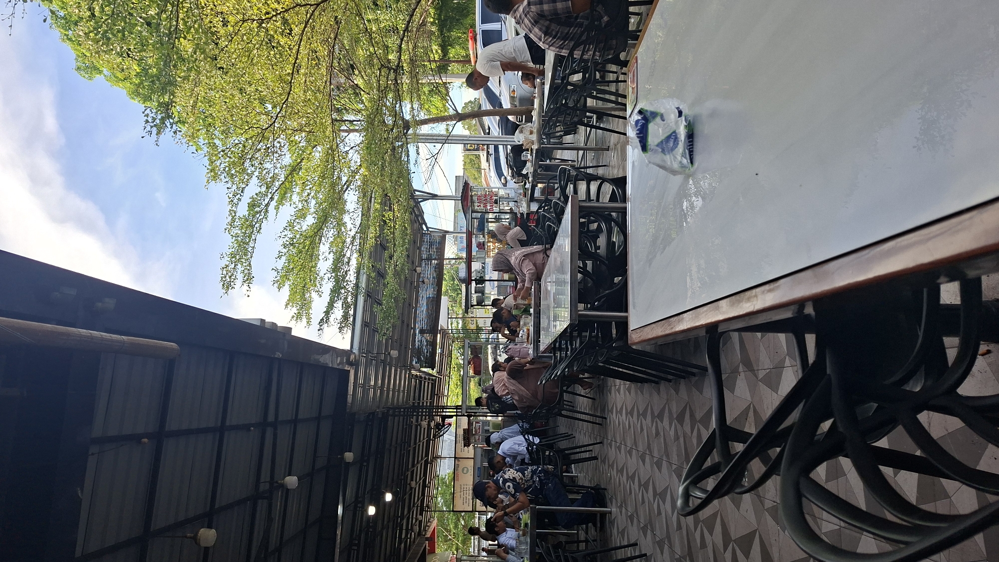 This is an outdoor scene at a restaurant in Keuniree, Indonesia, with tables and chairs set up under a covered roof. Several people are seated at tables, some eating, some conversing. A man in a white shirt is standing near a parked car, looking towards a table. Green trees are visible in the background, and the sky is partly cloudy. The flooring is tiled.