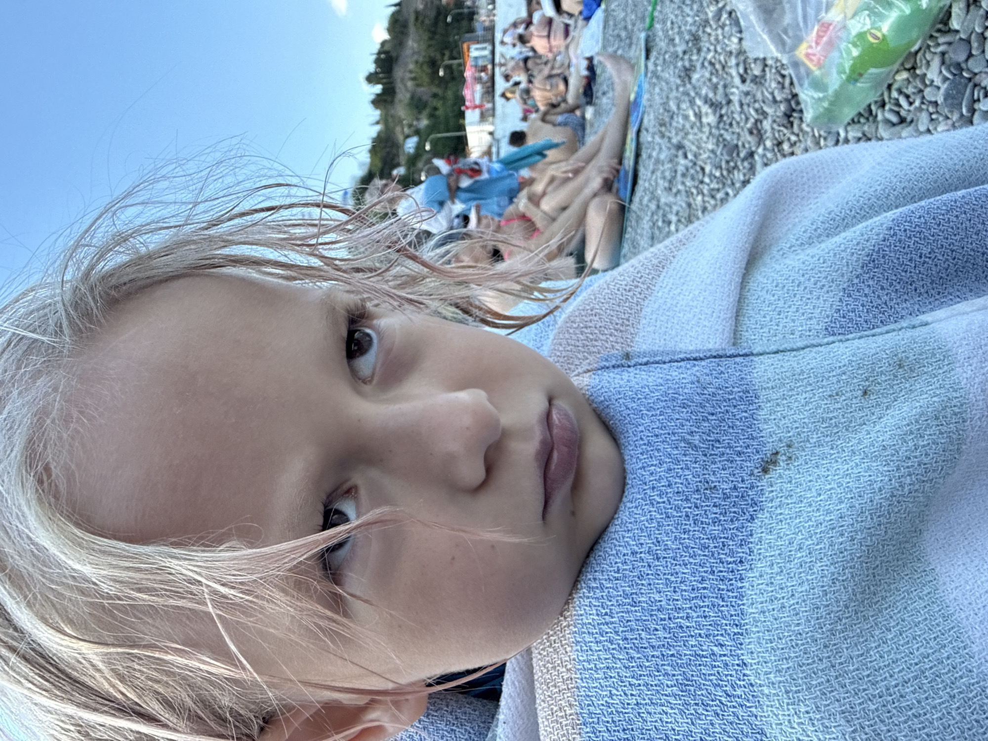 The media file is a close-up, selfie-style shot of a young child with blonde hair wrapped in a striped towel. The child's face is the primary focus, with their eyes looking slightly off-camera. The background is blurred, but appears to be a crowded beach scene with other people sunbathing and a hillside with trees and buildings. The sky is blue and clear, suggesting a sunny day. The overall impression is one of a casual, outdoor setting, likely a vacation or day trip to the beach. The child's expression is neutral. The time of day appears to be daytime, given the bright sunlight.